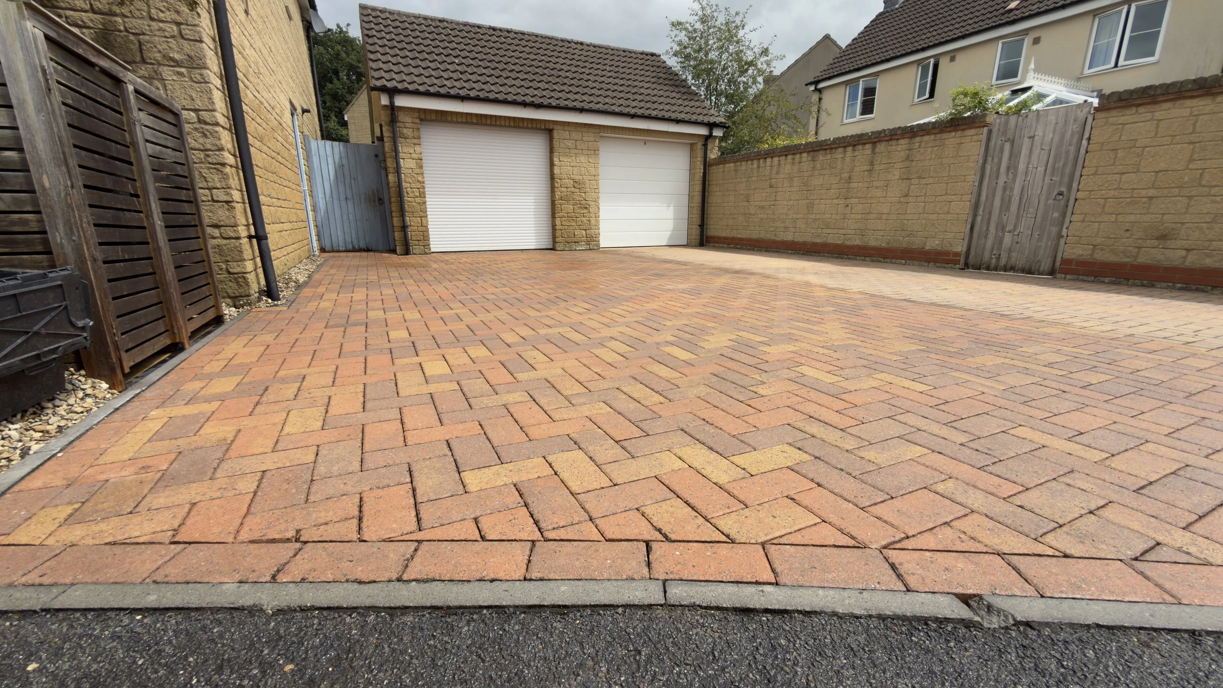 A paved driveway with red and yellow bricks, bordered by gravel and a black drainage grille. In the background, there are houses with brick and stucco exteriors, a small shed with a garage door, and wooden fences.