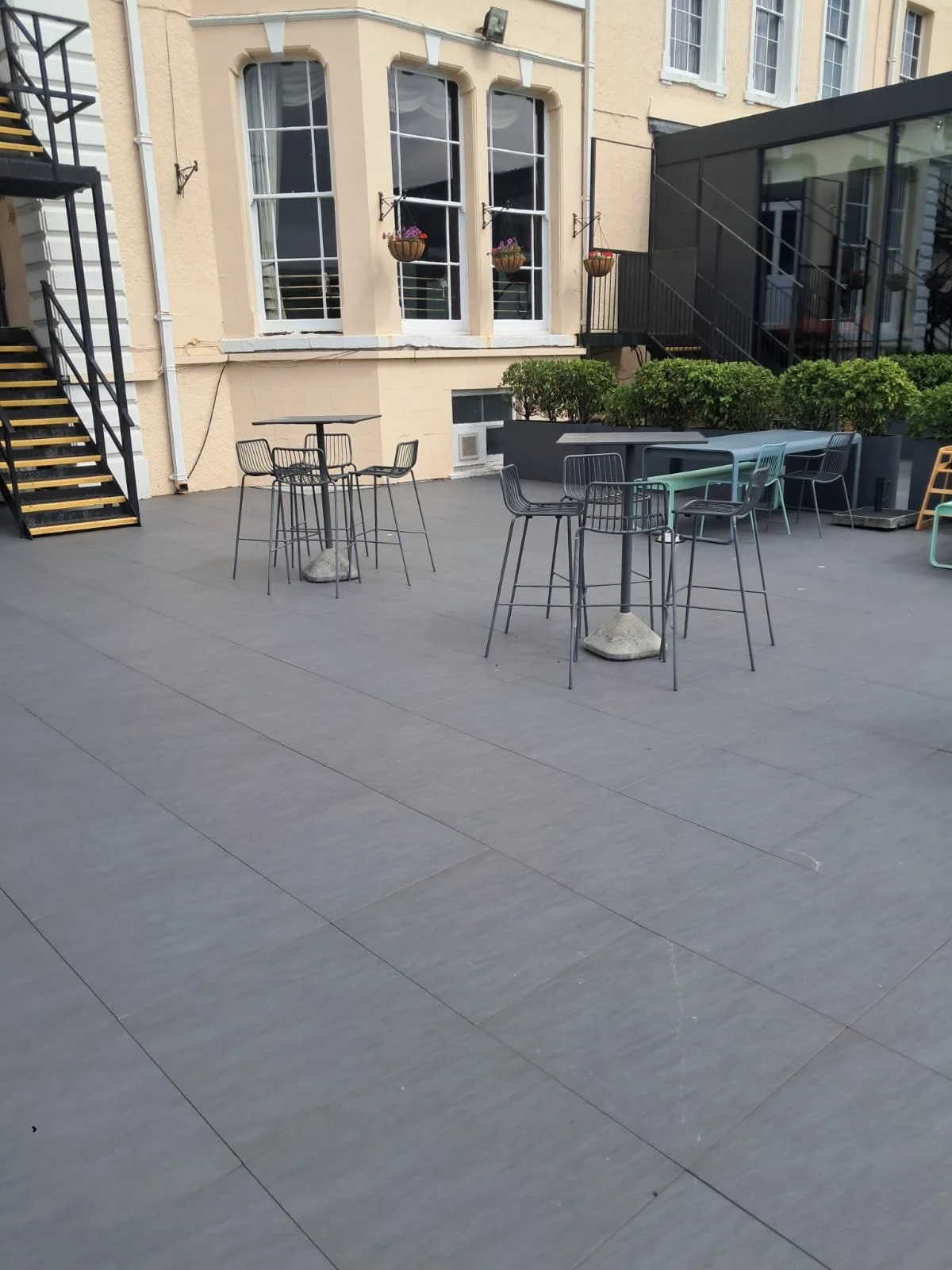 Empty outdoor patio with black wire chairs and small tables on a gray deck, next to a beige building with large windows and hanging flower baskets.