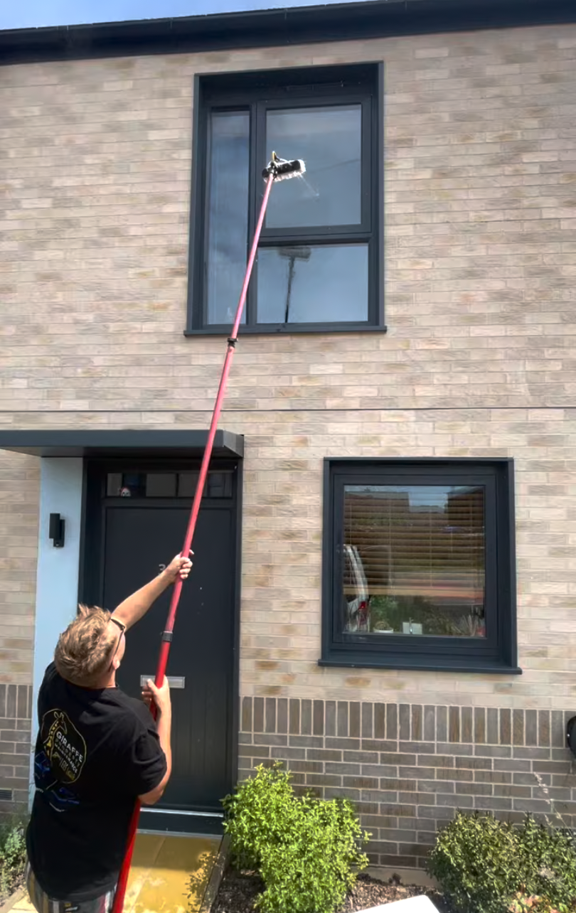 Person using a long cleaning tool to wash the exterior window of a two-story brick house.
