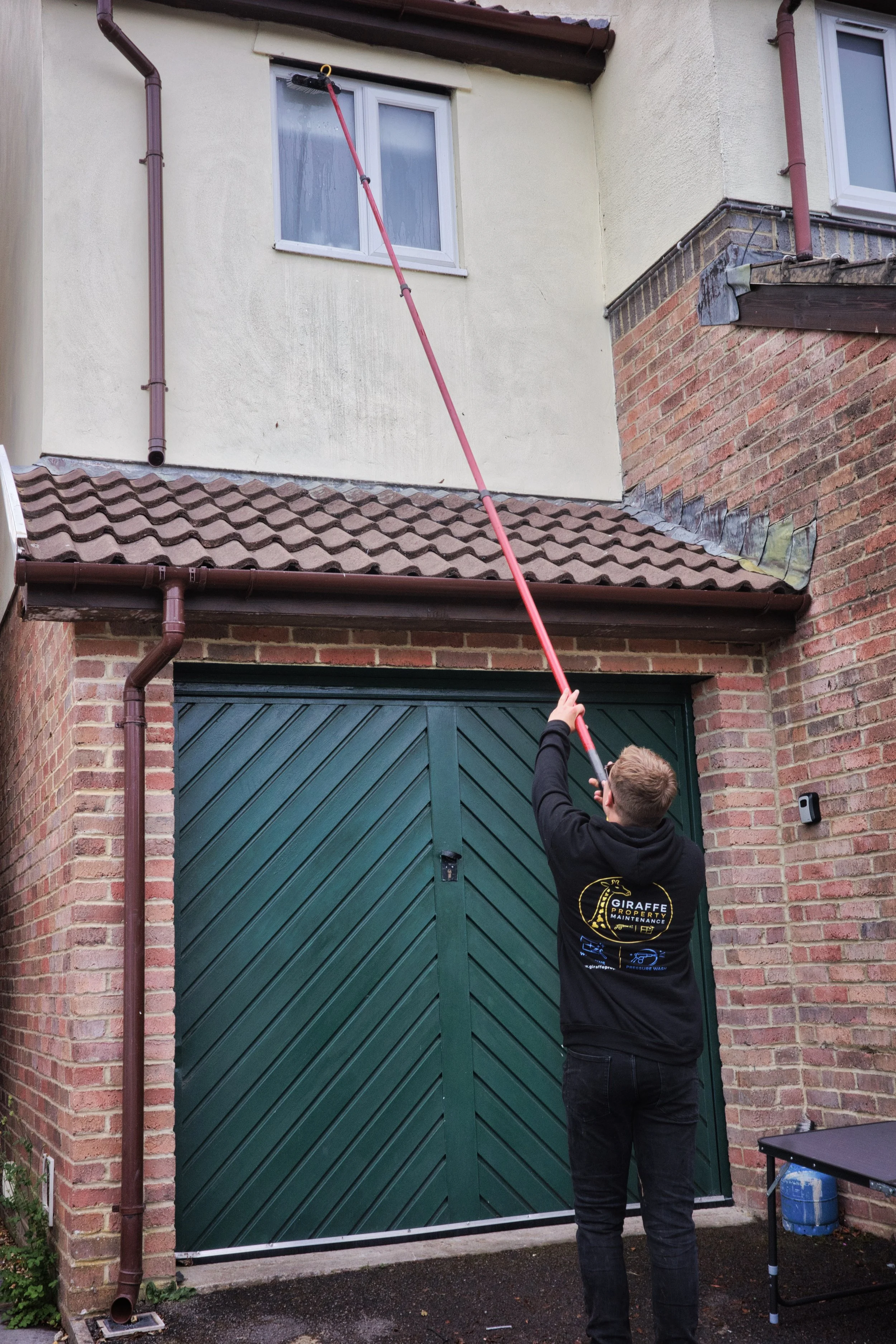 A person using a long pole to clean or wash the upper part of a building's exterior wall near a window and a gutter.