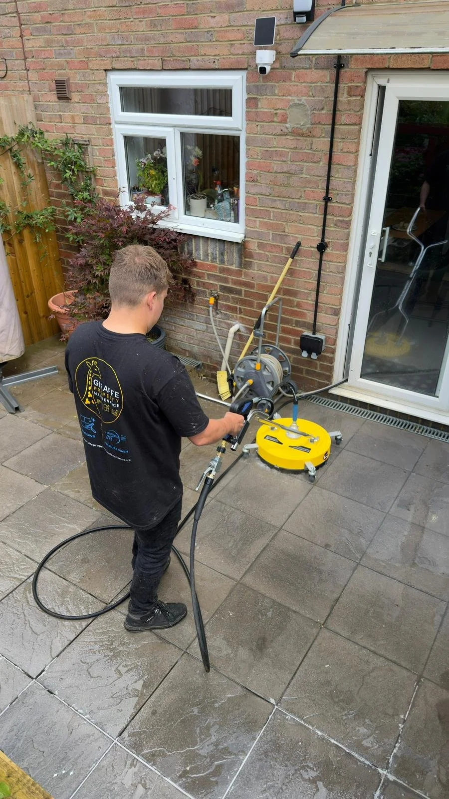 A person cleaning outdoor patio tiles with a pressure washer. The person is wearing a black T-shirt and black pants, standing on wet, gray stone tiles next to a brick house with a window and a sliding glass door.