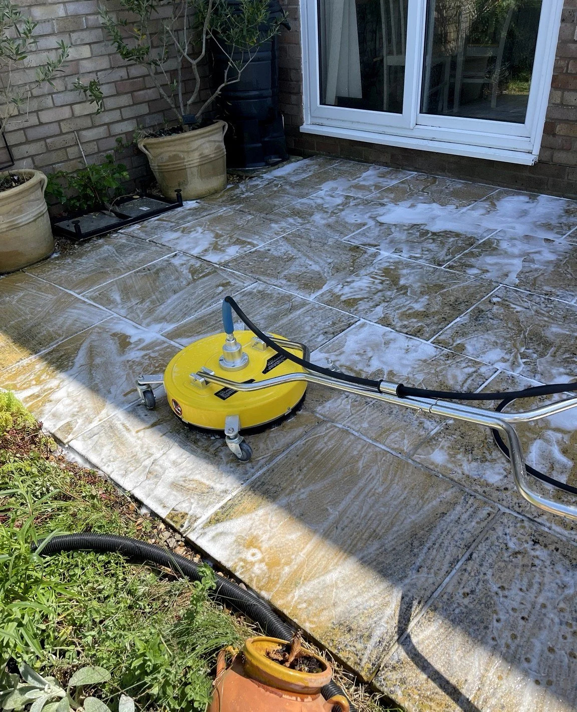 A pressure washer cleaning a stone patio outside a house with potted plants along the wall and a sliding glass door.