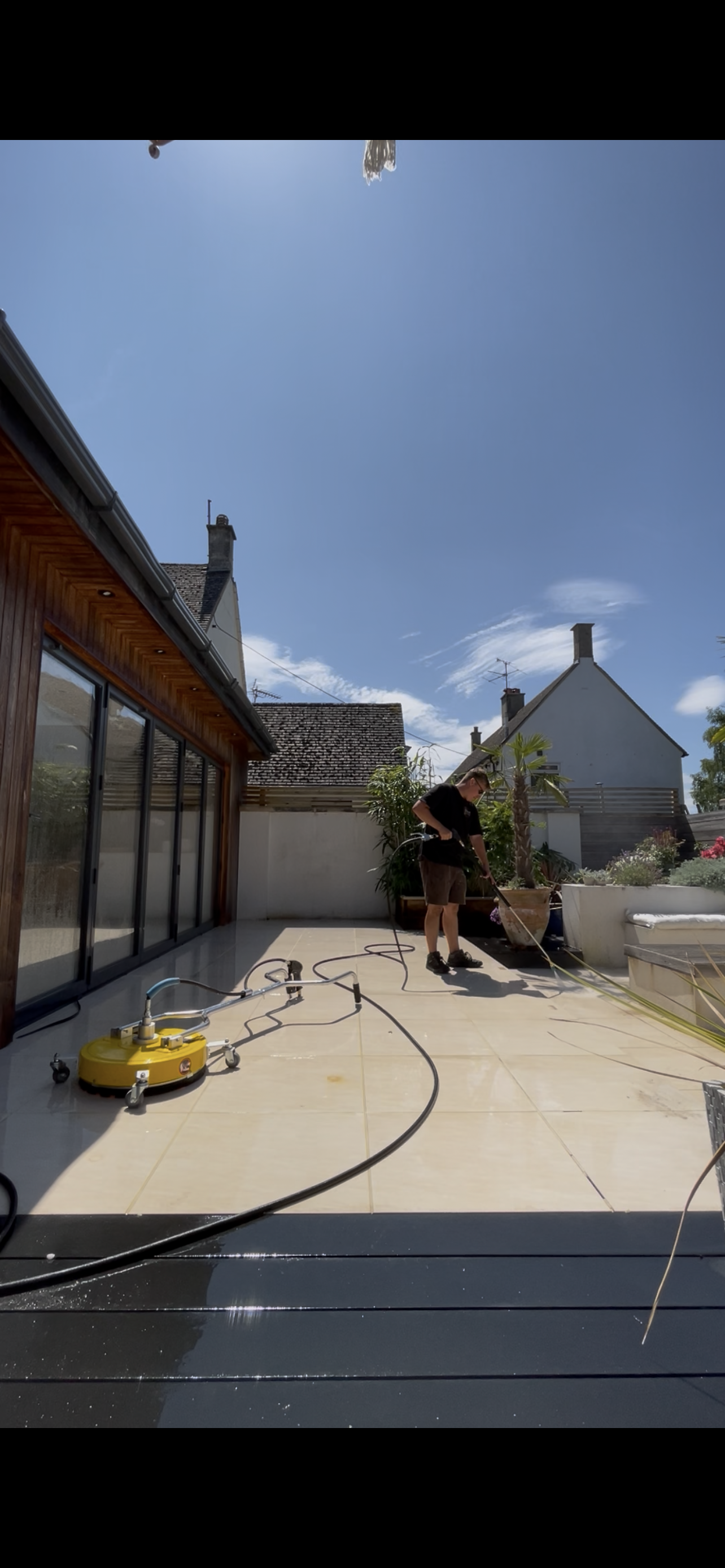 A man cleaning an outdoor patio using a self-propelled pressure washer on a bright, sunny day. The patio has beige tiles, large glass doors, and houseplants in the background.