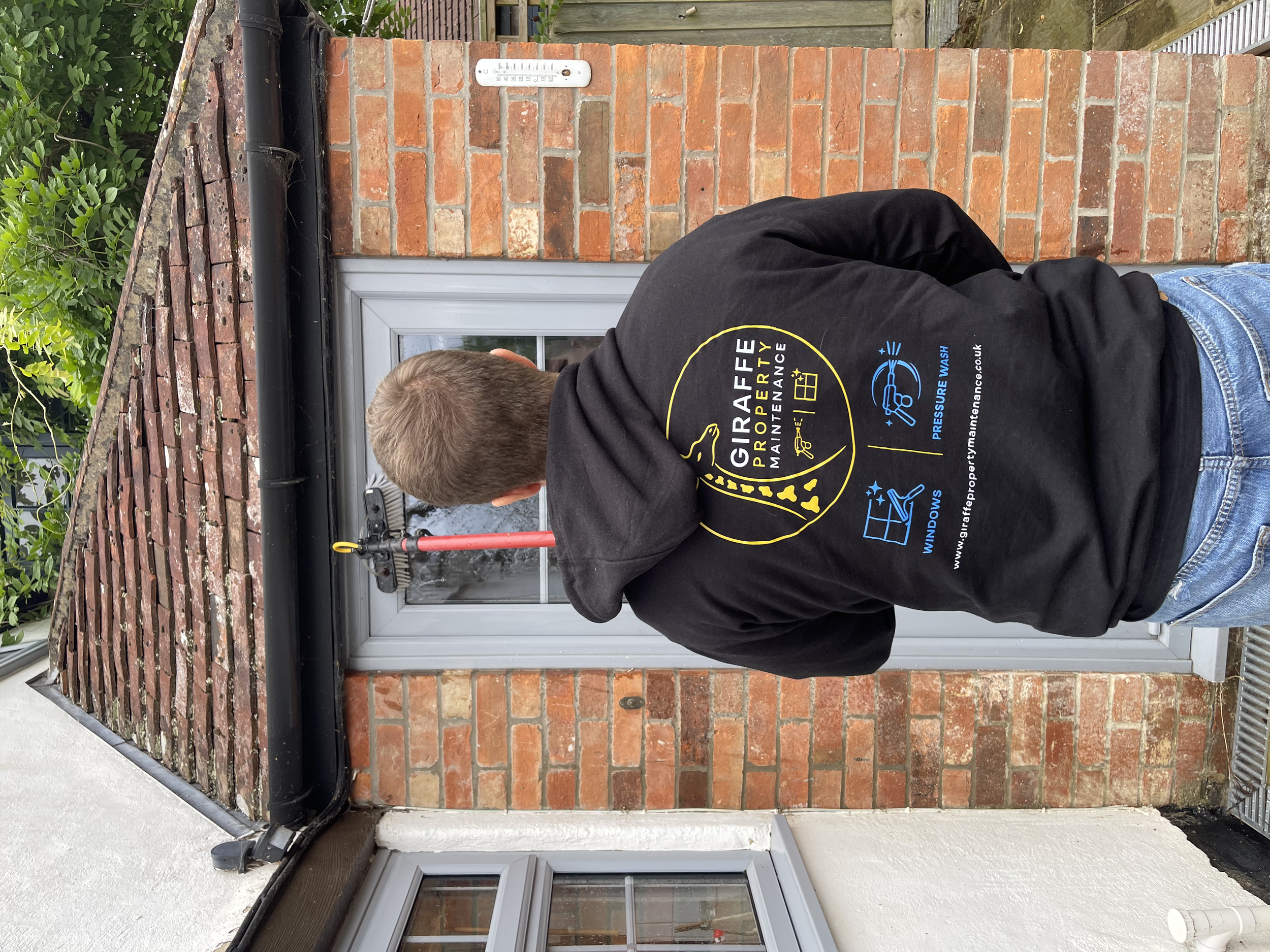 A man with short blonde hair wearing a black Giraffe Property Maintenance T-shirt is working on a brick wall near a white door. The wall has a thermometer attached, and there is greenery and a wooden fence in the background.