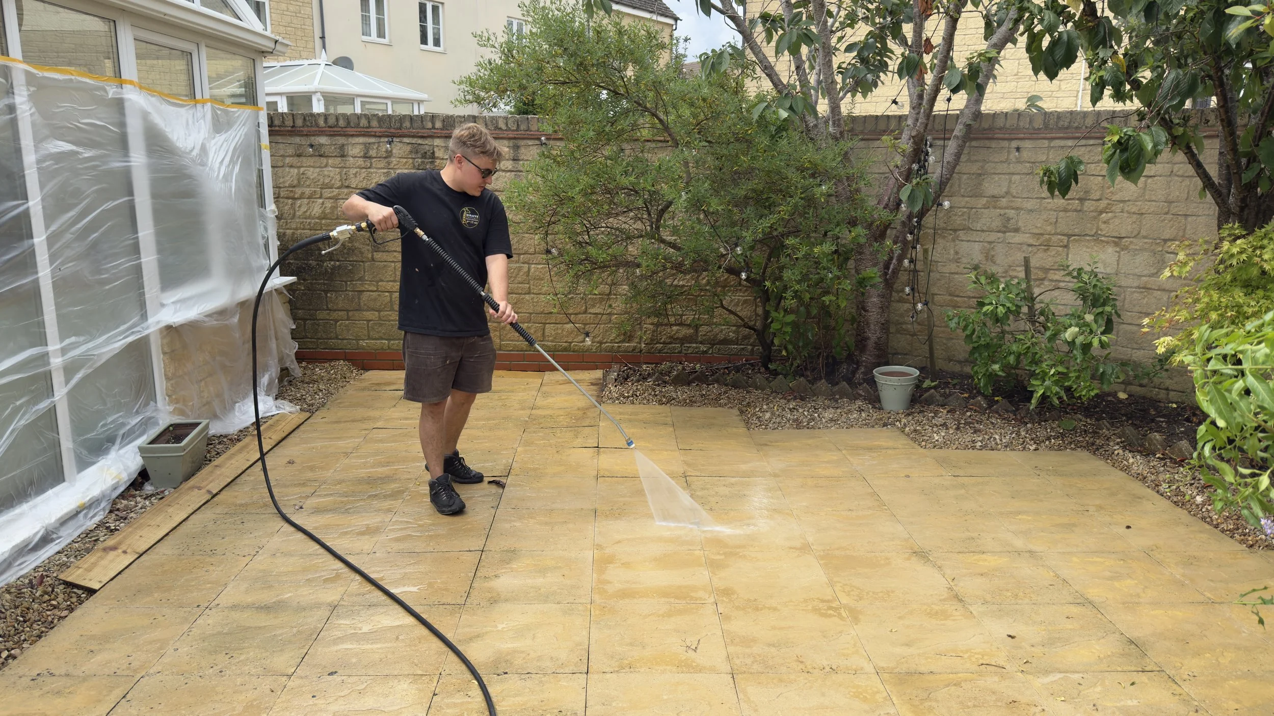 A young man wearing a black t-shirt, shorts, and sunglasses is pressure washing a wooden patio in a backyard. There are trees, potted plants, and a brick wall in the background, along with part of a house with large windows and a conservatory.