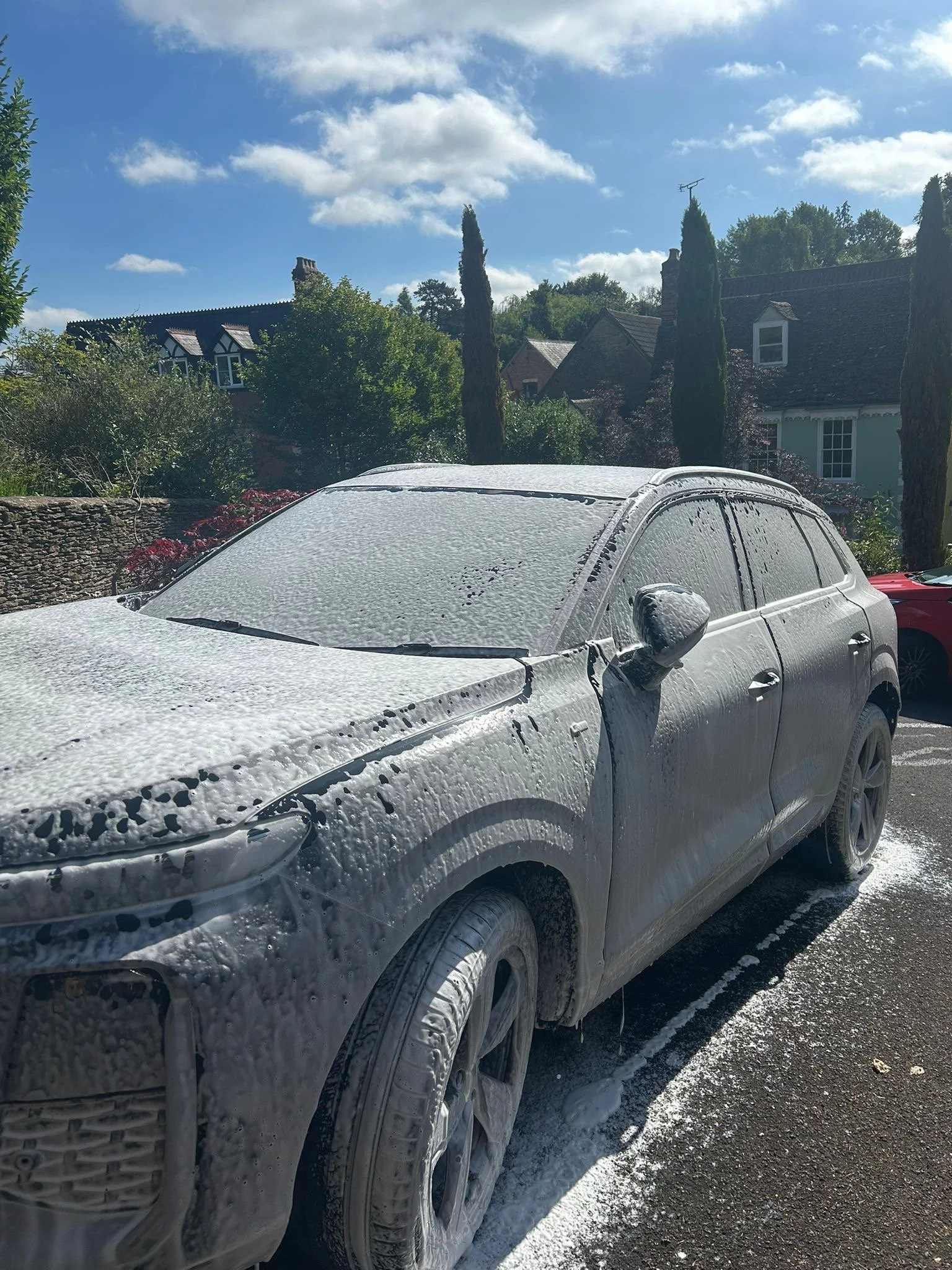 A white car covered in soap bubbles parked outside houses on a sunny day.