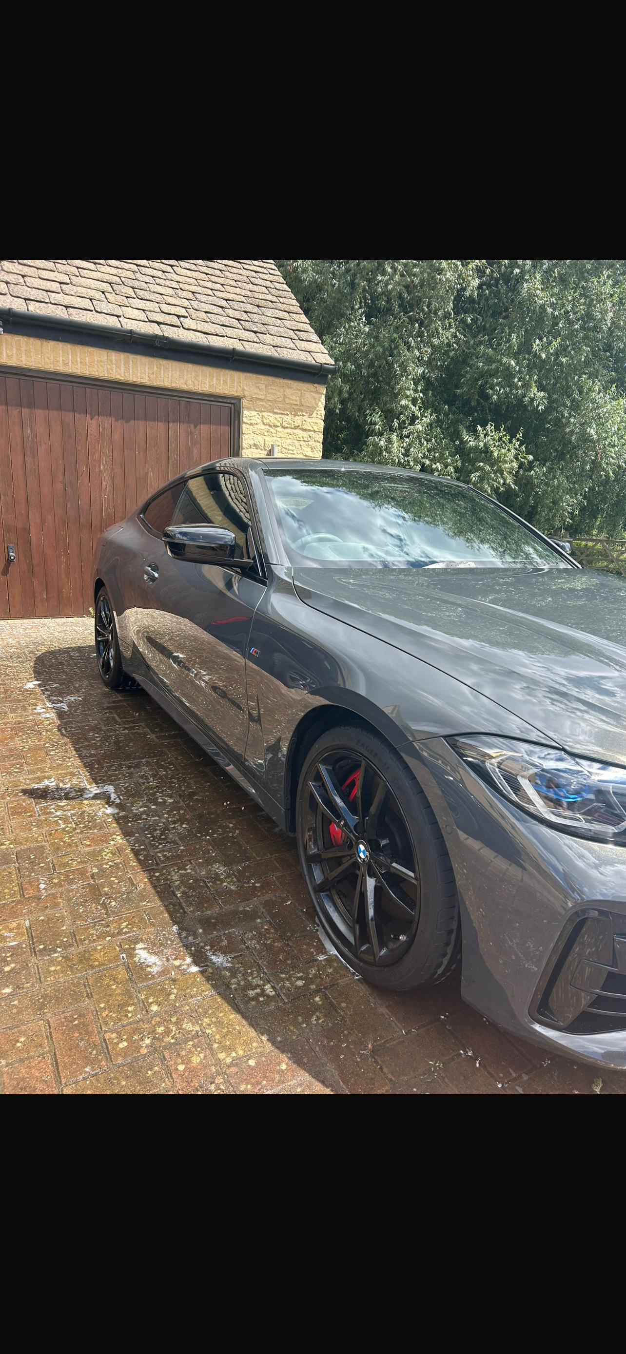 A gray BMW M Series coupe with black wheels and red brake calipers parked on a brick driveway in front of a wooden garage door at a house surrounded by green trees.