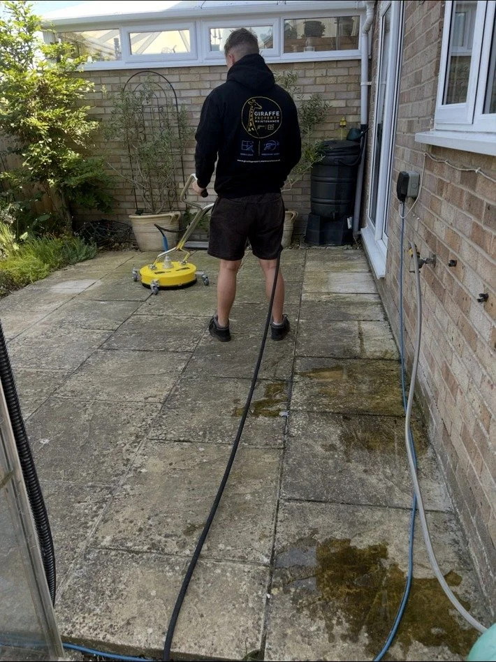 Person cleaning moss off patio tiles using a yellow pressure washer, wearing a black hoodie and black shorts, in a backyard with brick walls and garden plants.