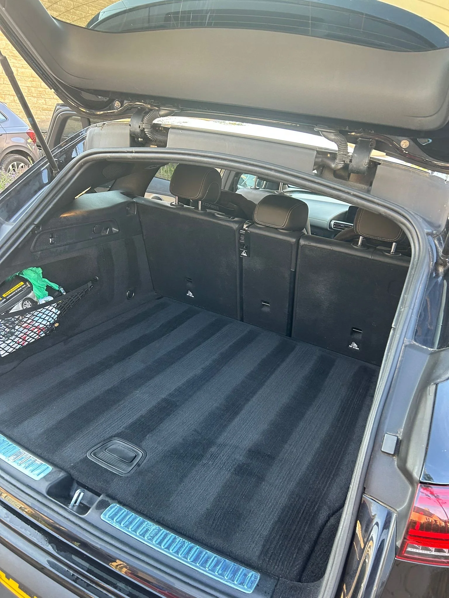 The photo shows the open trunk of a car, revealing a black carpeted interior with some small items like a green bag with yellow and black markings and a black mesh pocket on the side. The rear seats are visible with headrests, and the rear window is 
