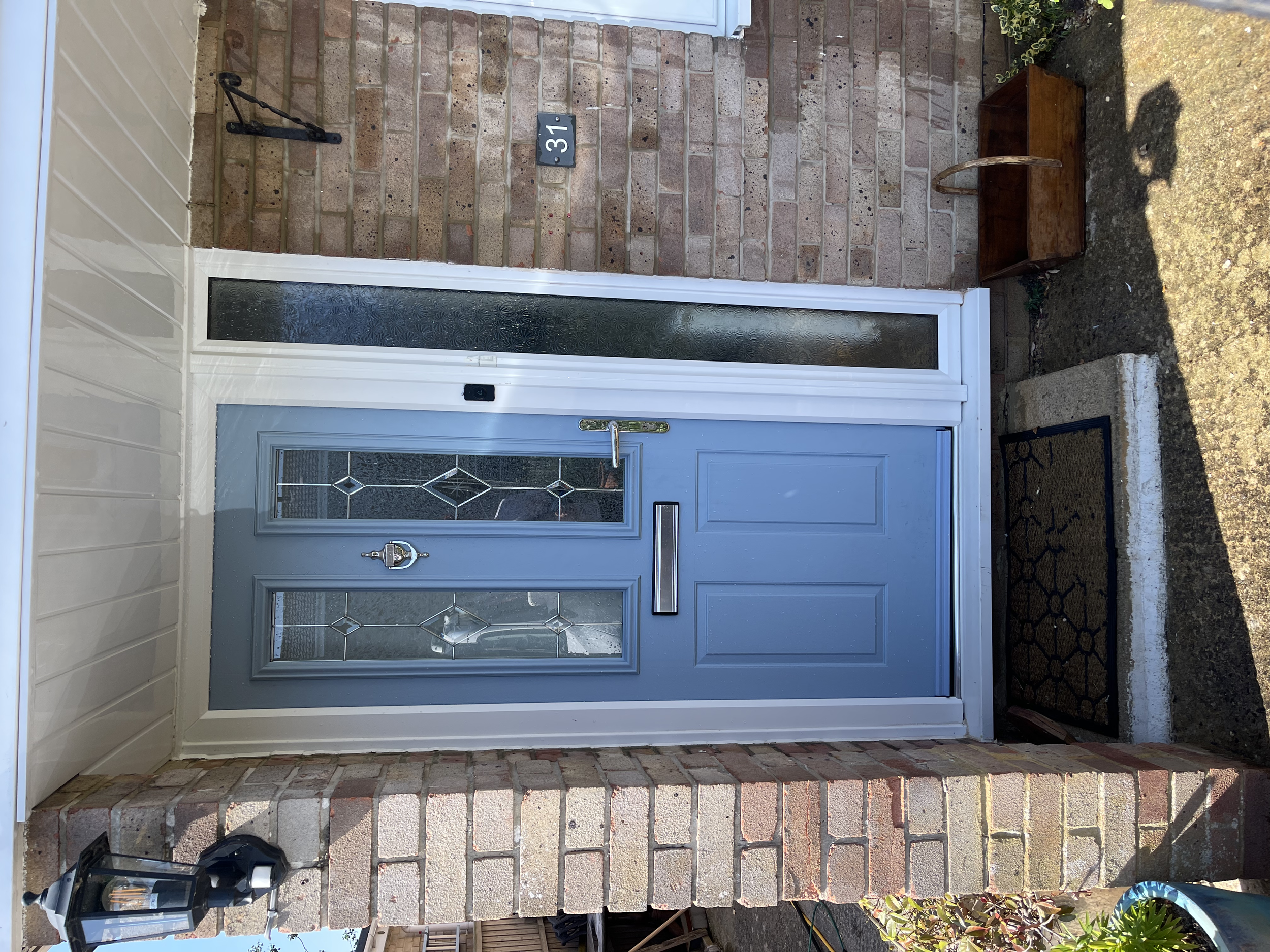 The front door of a house with a brick wall, house number 31, a glass panel door, and an outdoor lantern. There is a small wooden table with a curved leg and landscaping on the right side.