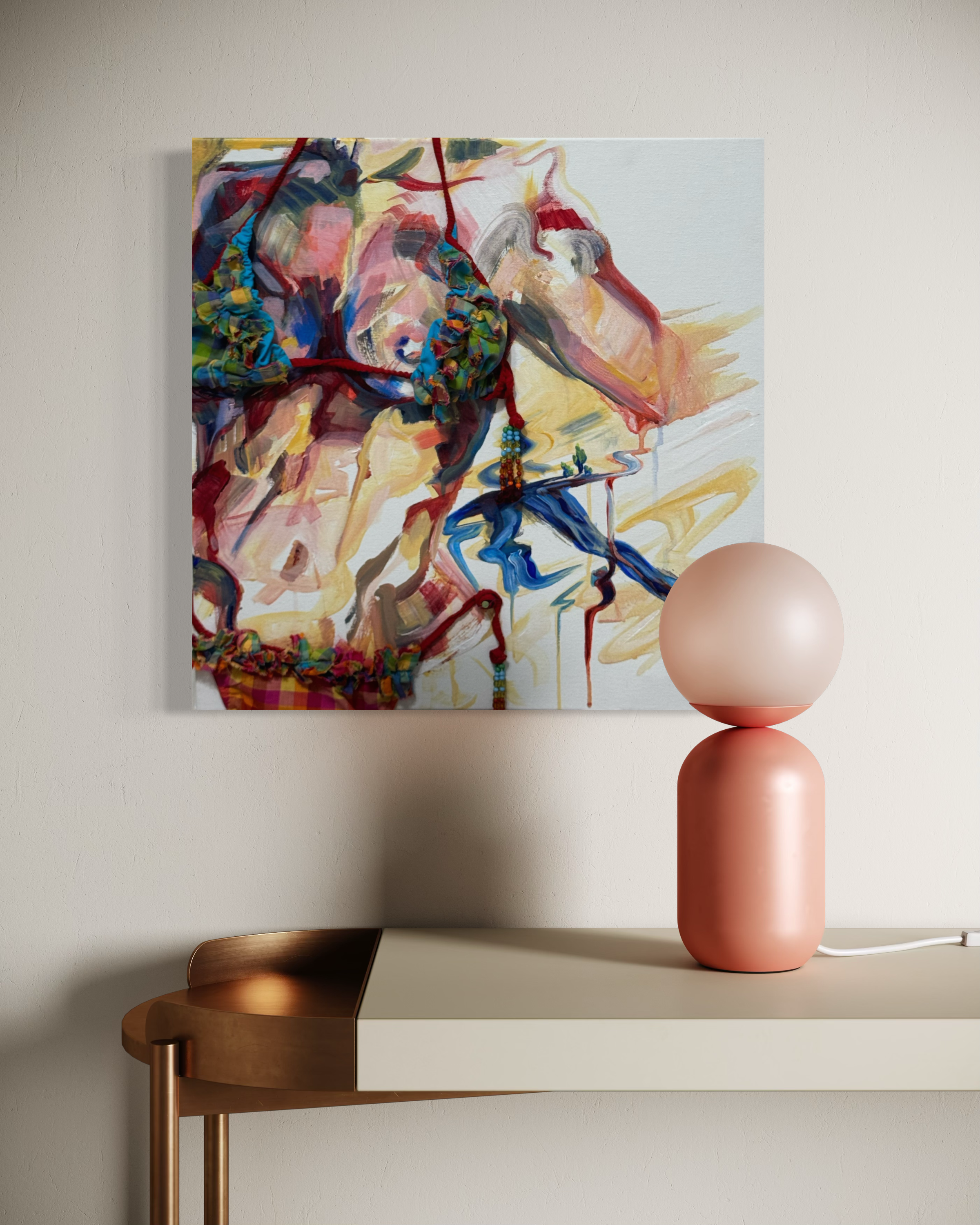 An abstract painting on a canvas features colorful beads and fabric strands draped across the surface. The painting is mounted on a light-colored wall above a modern table with a metallic, copper-colored edge, next to a pink ceramic lamp with a rounded base.