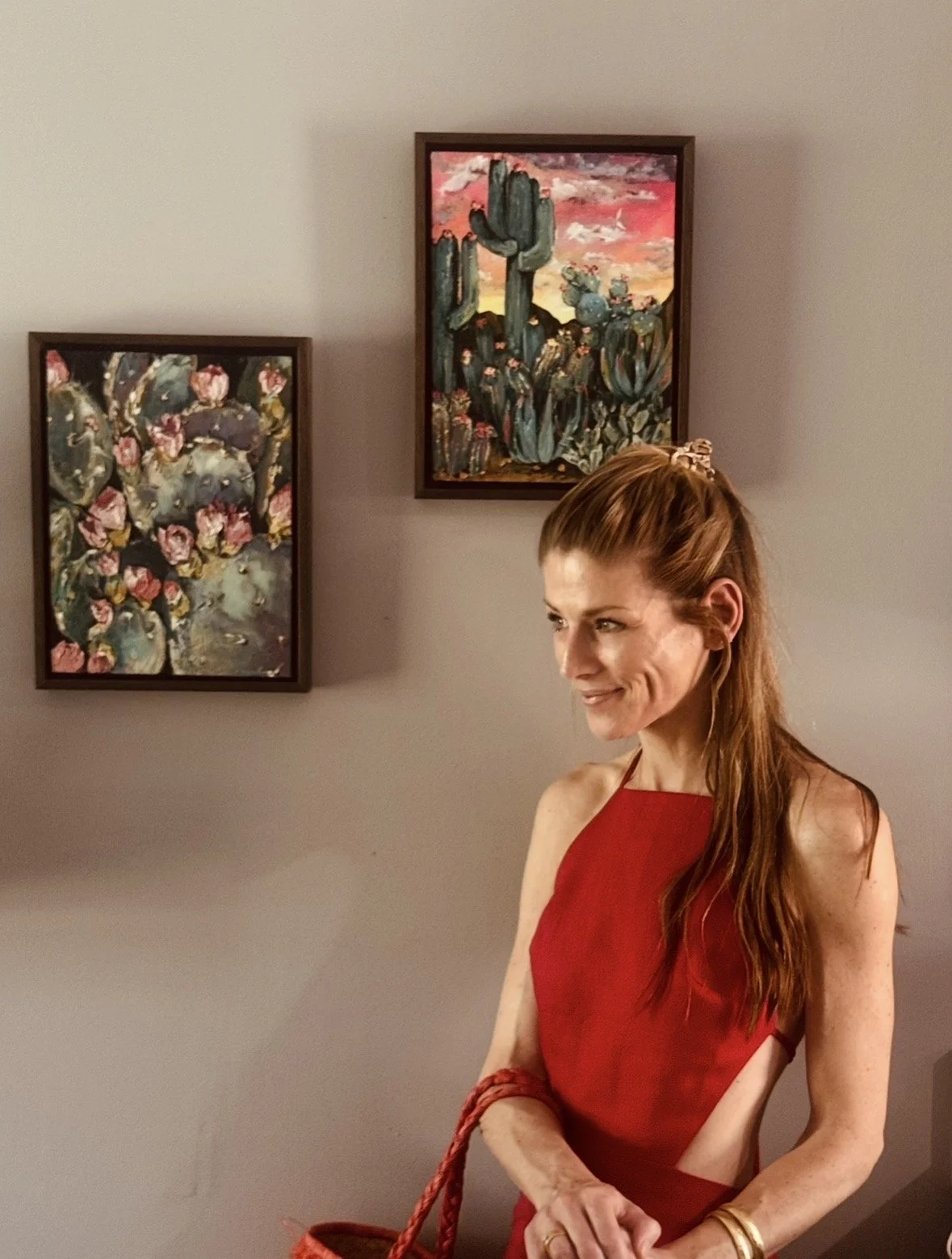 A woman with red hair wearing a red dress standing in front of a wall with three paintings of cacti and succulents.