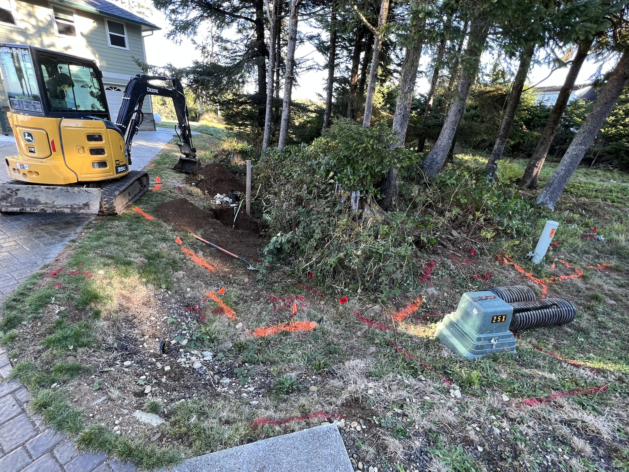 A small yellow John Deere excavator digging a trench in the yard of a house with trees and shrubs nearby. The trench is marked with orange spray paint and small orange flags.