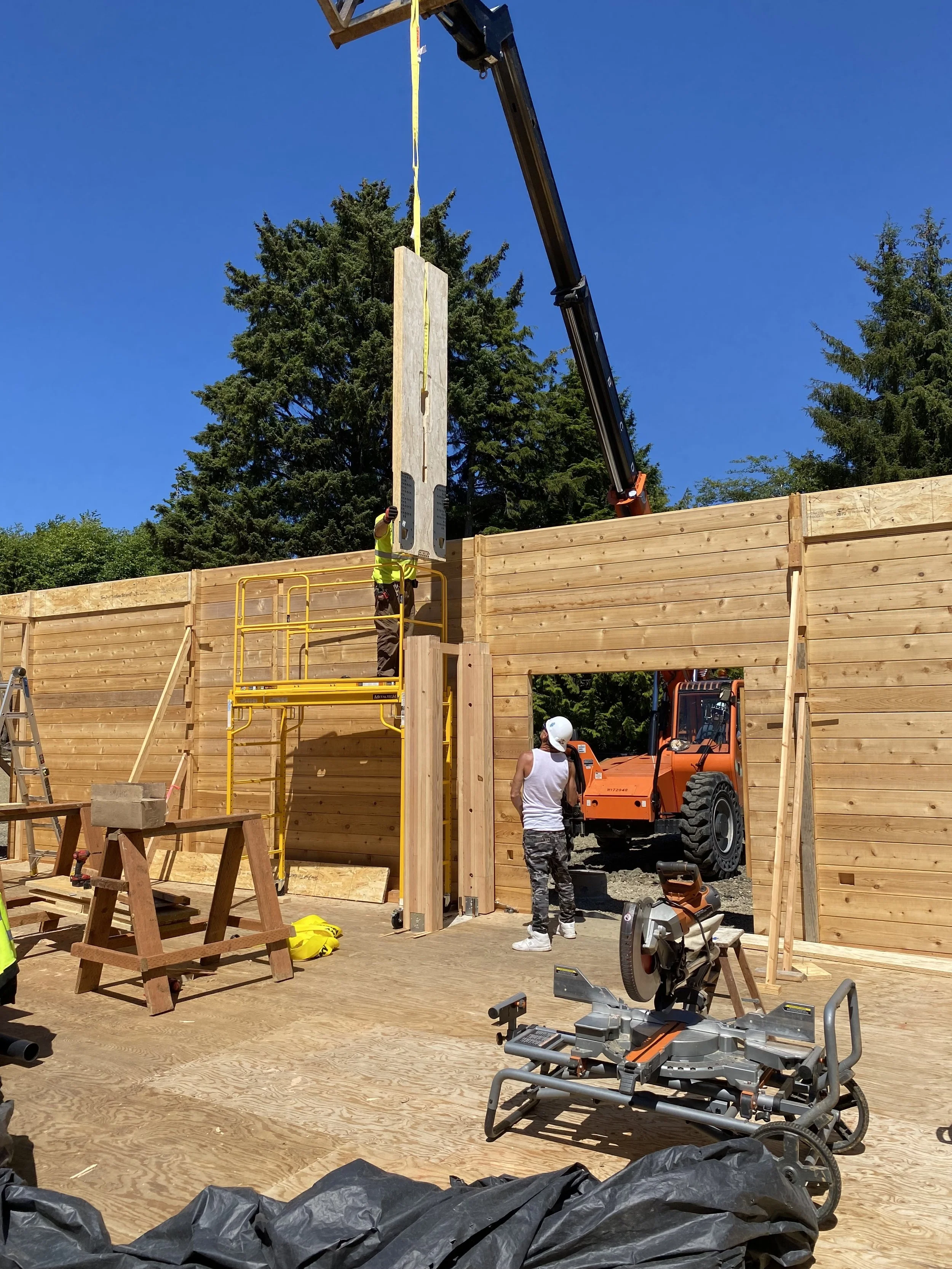Construction site with workers installing a large wooden wall panel using a crane. One worker is operating the crane while another stands on the ground observing. There is construction equipment, tools, and safety gear visible, with a background of t
