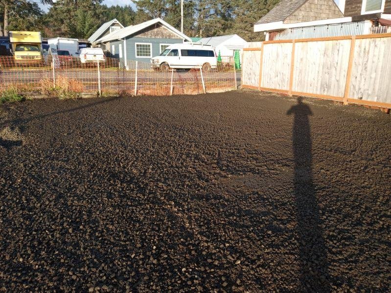 A freshly tilled garden or yard area with dark, loose soil, enclosed by orange plastic fencing and a wooden fence, with houses and parked vehicles in the background.