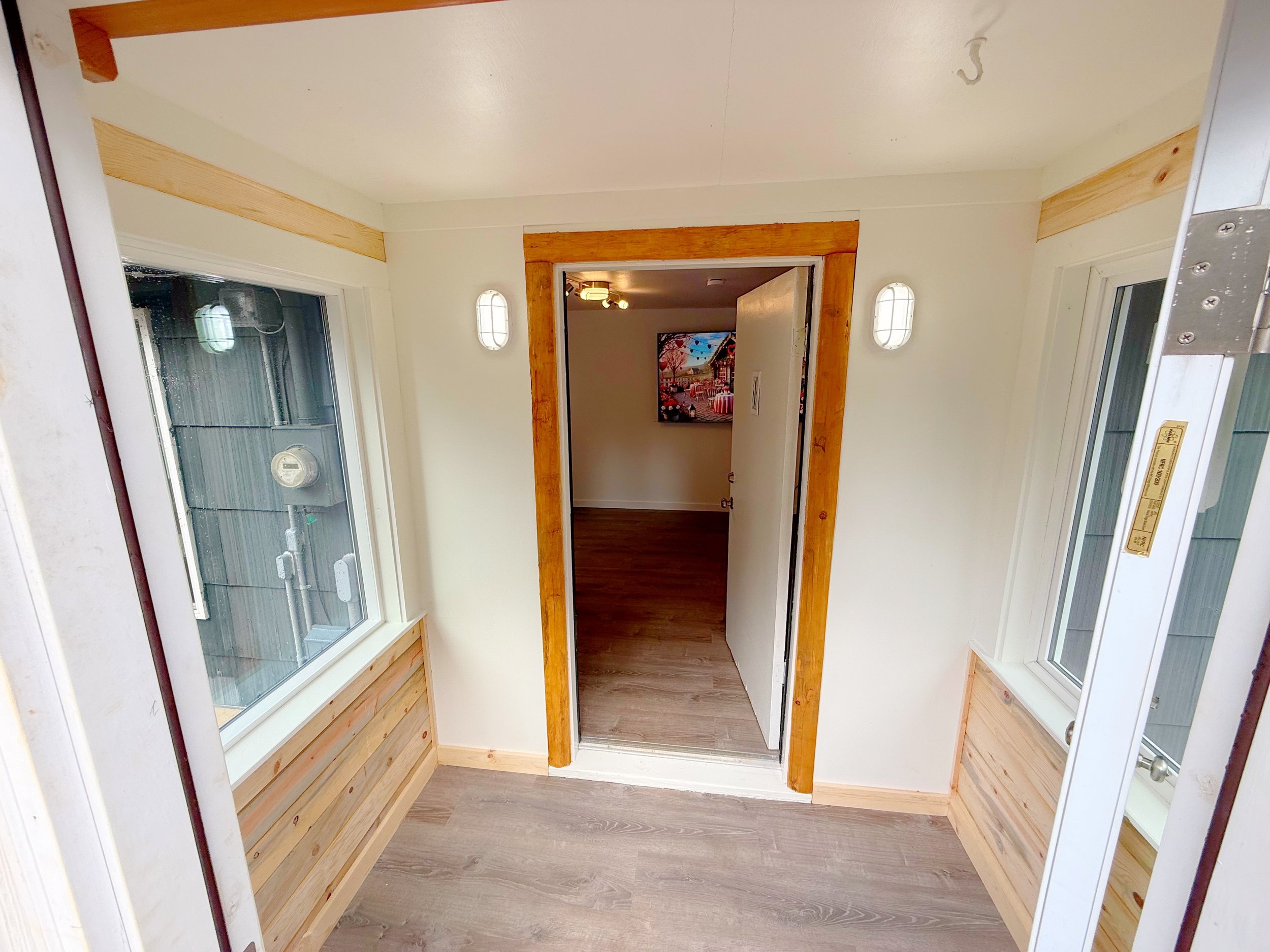 An indoor room with white walls, wooden trim, and a wooden floor. There are two large windows on each side and a doorway at the far end leading to another room with colorful wall decorations. The room is well-lit with ceiling and wall lights.