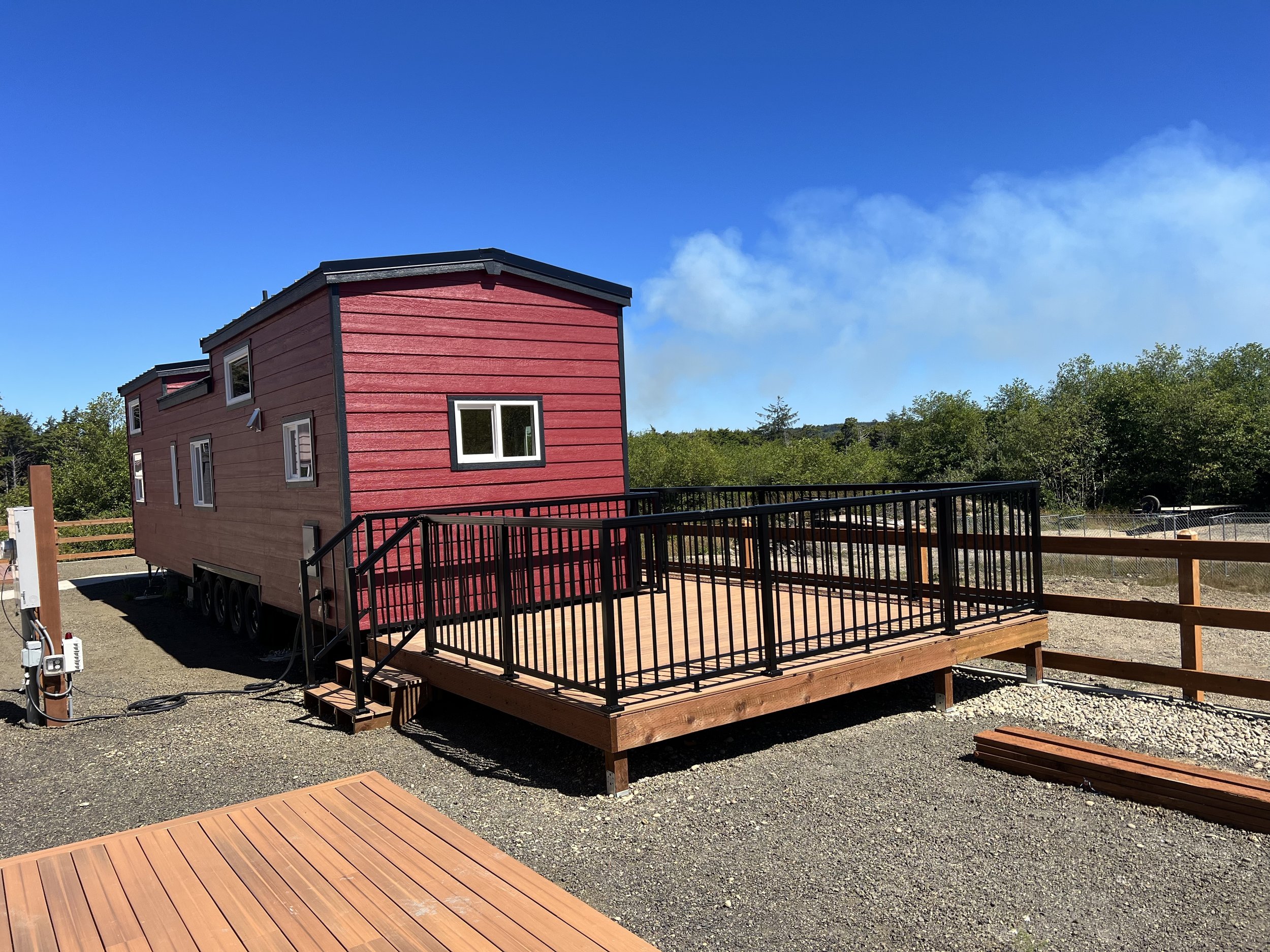 A residential tiny house on wheels painted red with black trim, situated on a gravel lot with nearby wooden deck and fencing, under a partly cloudy blue sky with trees in the background.