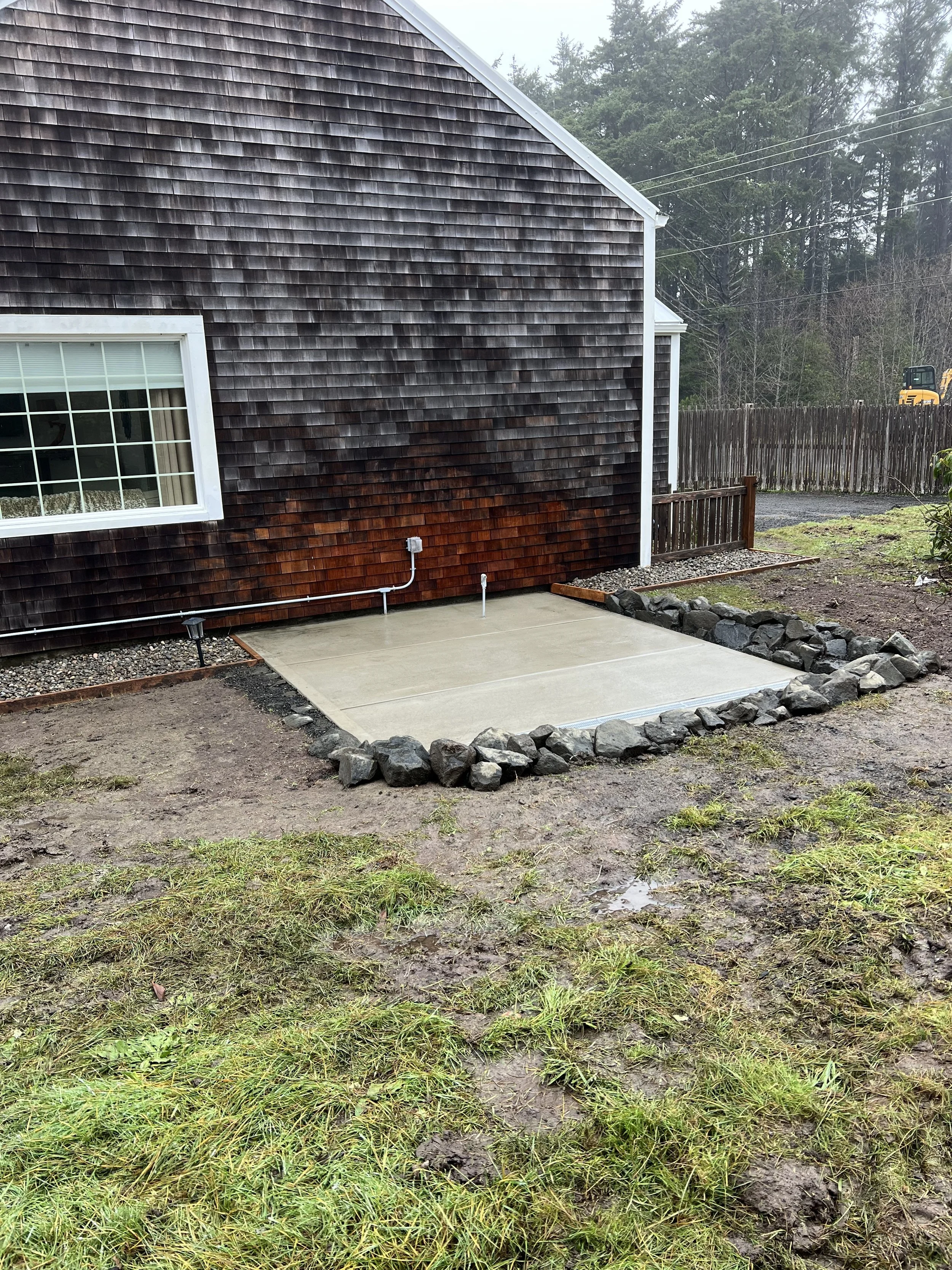 A backyard with a deck, a gardening bed with rocks, a muddy lawn with grass, a wooden fence, and trees in the background on a cloudy day.