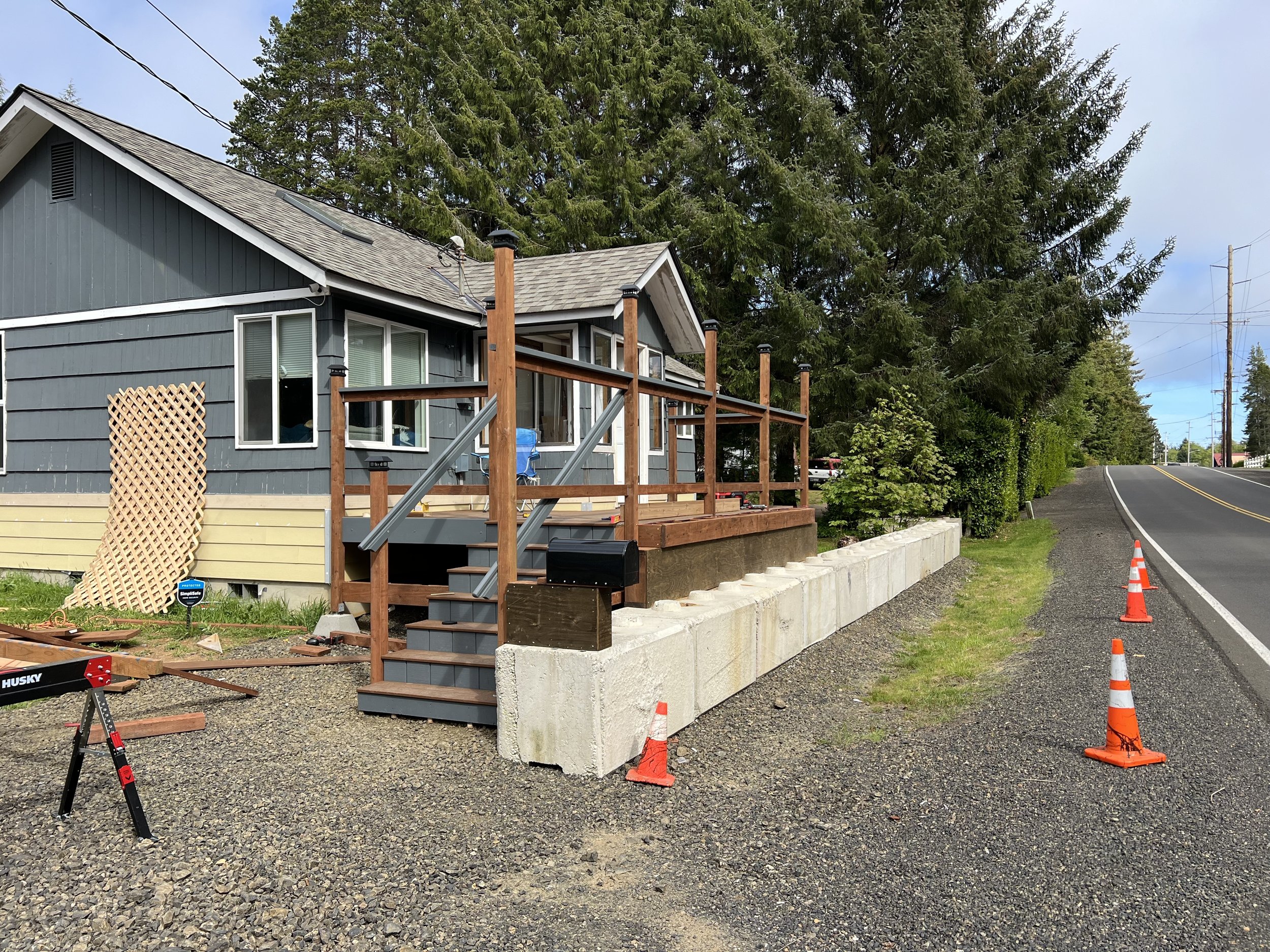 A house under construction next to a gravel road with orange construction cones, surrounded by trees and power lines.