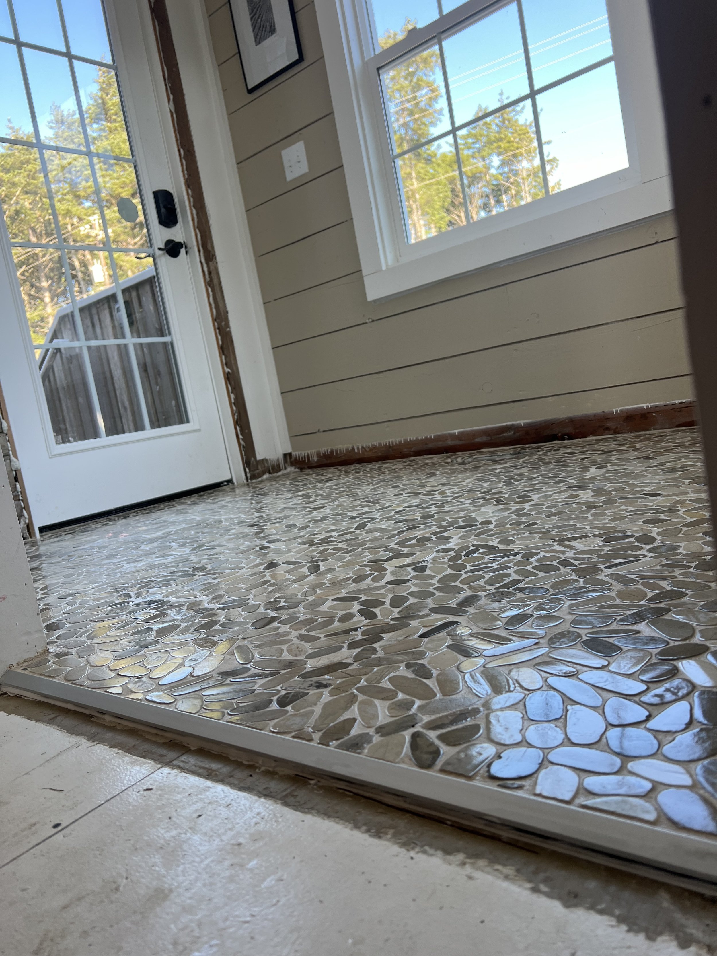 Interior view of a room with a glass door and large window, beige horizontal paneling on the walls, and a floor with mosaic tile pattern of pebble-like shapes in earthy tones. Outside trees and a bright blue sky are visible through the window and doo