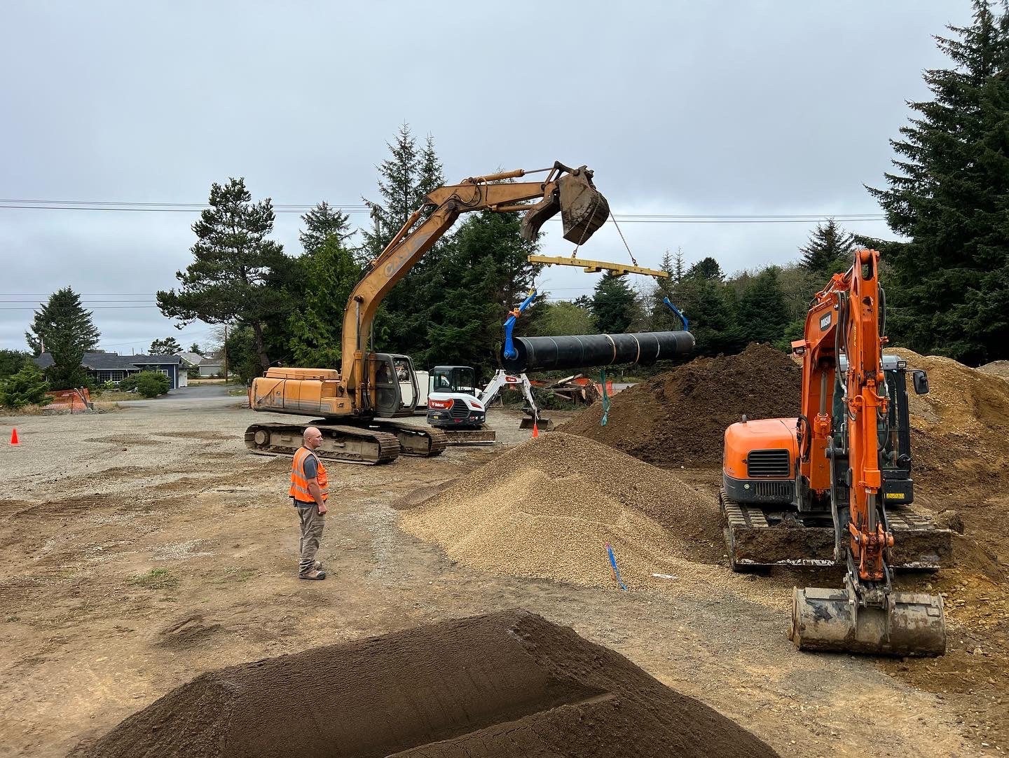 Construction site with excavator lifting a large pipe using a crane, and a worker in an orange safety vest observing on a cloudy day.