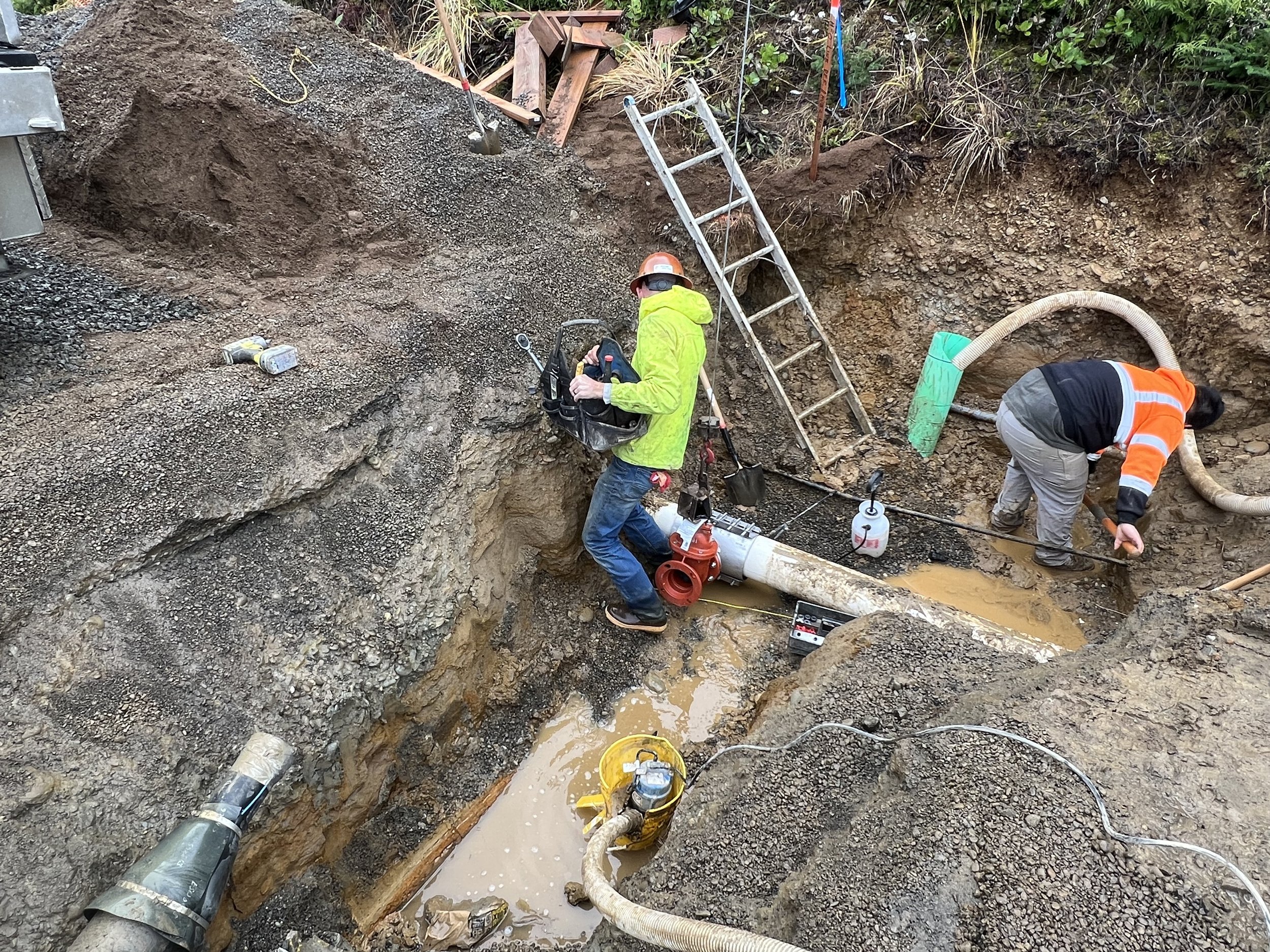Workers are repairing underground pipes in a large trench. One worker is operating equipment in the trench, and another worker is handling hoses and tools. The trench contains water, and PVC pipes are visible on the ground.
