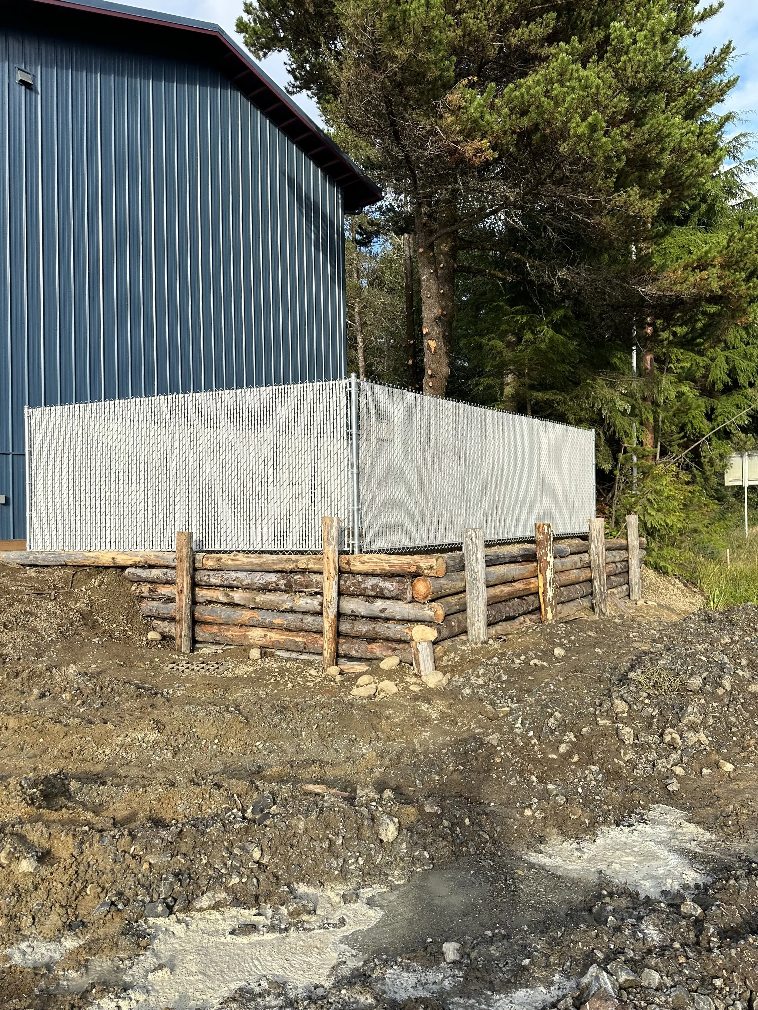 A construction site with dirt and rocks in the foreground, a green metal building on the left, a chain link fence, and large trees in the background.