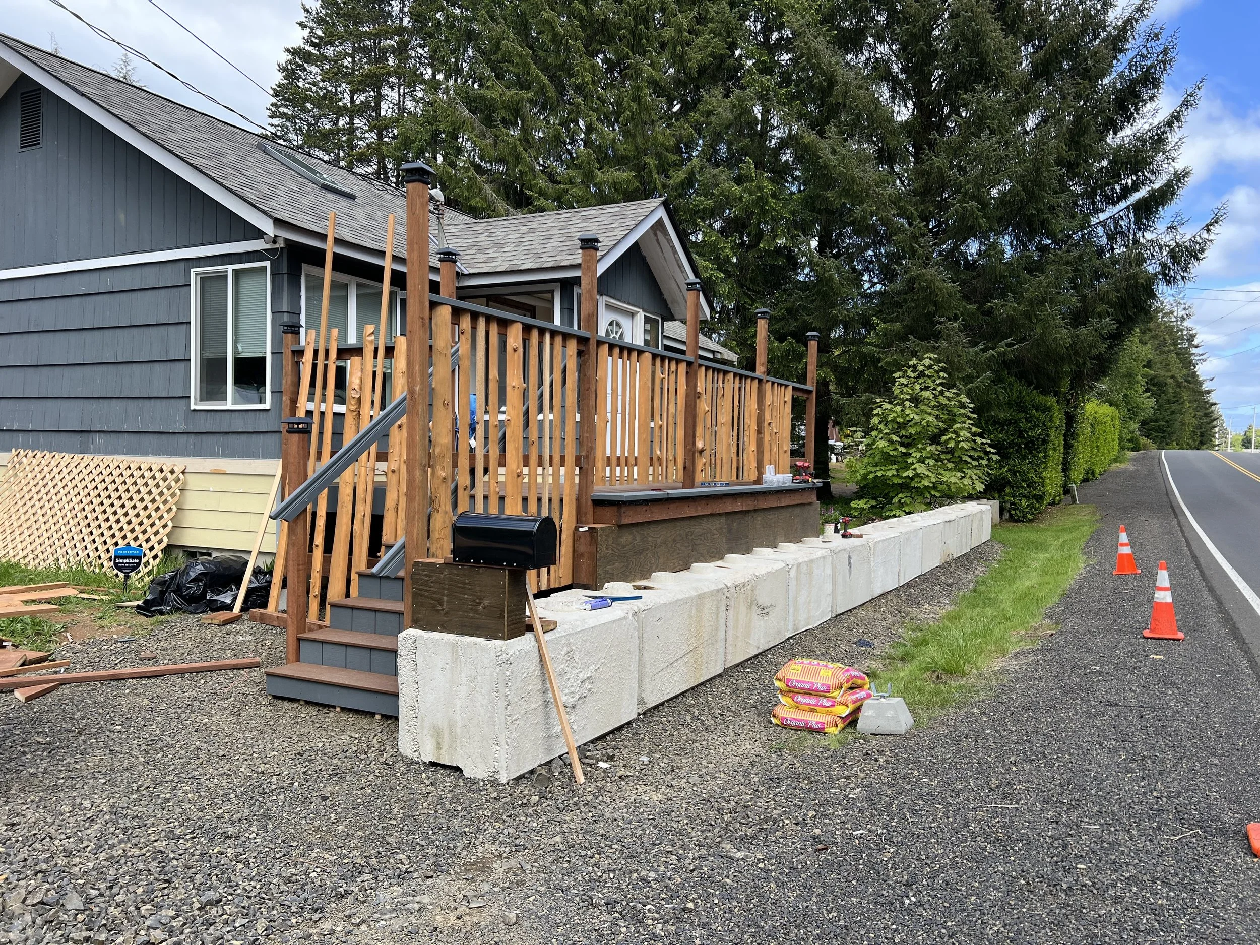 A house with gray siding and a wooden deck under construction, with construction materials and tools on the ground, and orange safety cones along the side of a rural road.