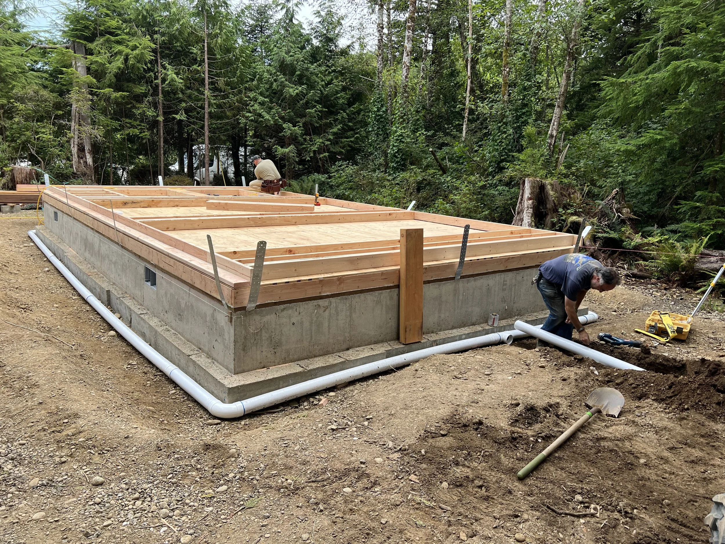 Construction workers building the wooden frame of a small structure on a concrete foundation in a forested area.