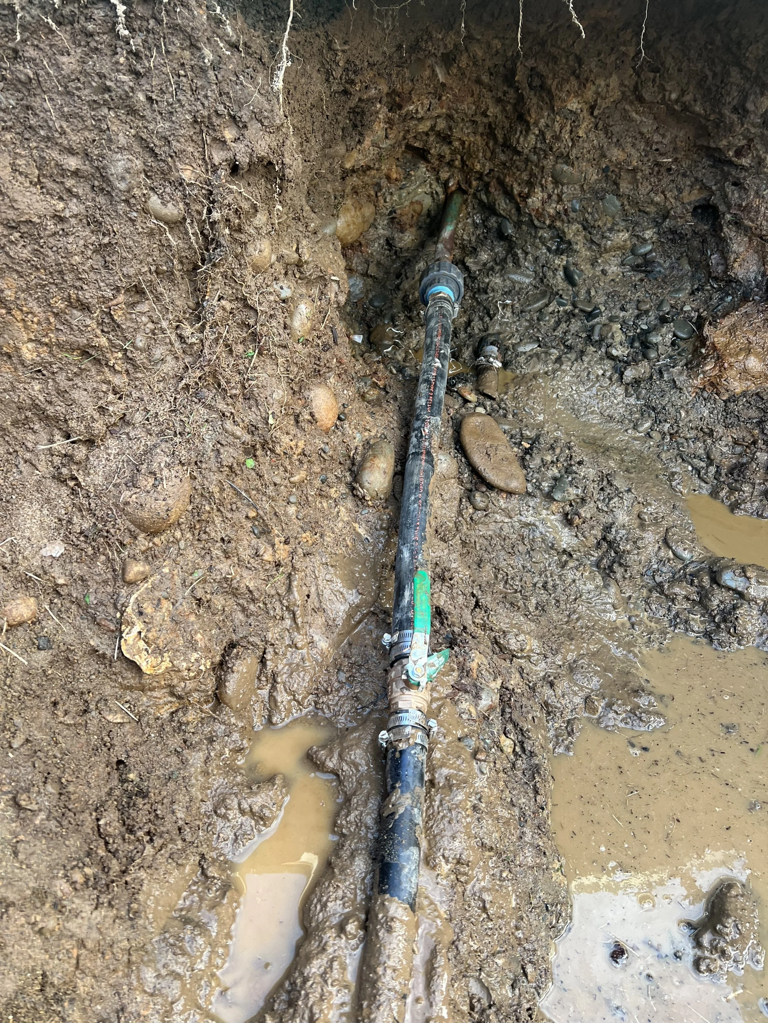 A muddy construction site with a pipe running through the dirt and rocks, surrounded by wet, loose soil and small water pools.