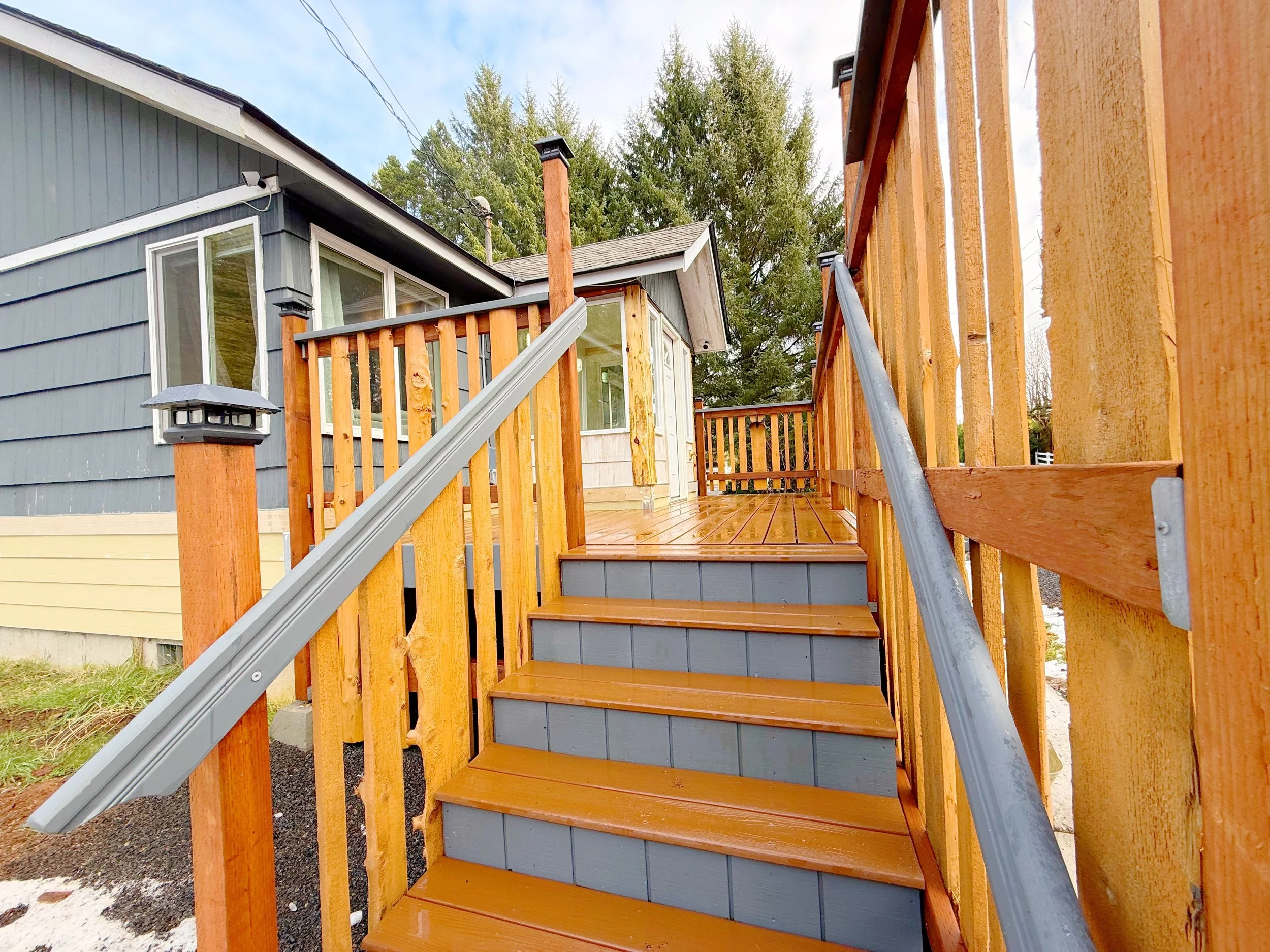 Unfinished wooden deck with stairs leading to the ground, attached to a gray house with large windows, decorated with black outdoor lights, surrounded by trees and blue sky.