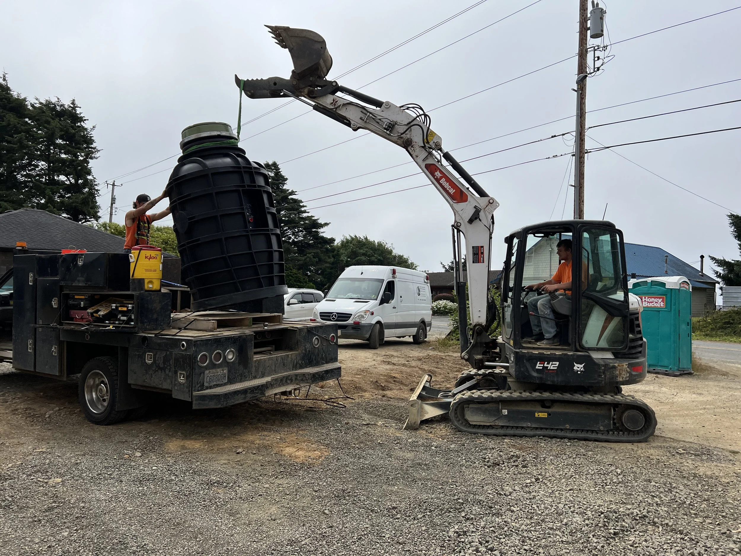 A construction site with a person operating a small excavator, lifting a large plastic container with a robotic arm, while another person is observing nearby.