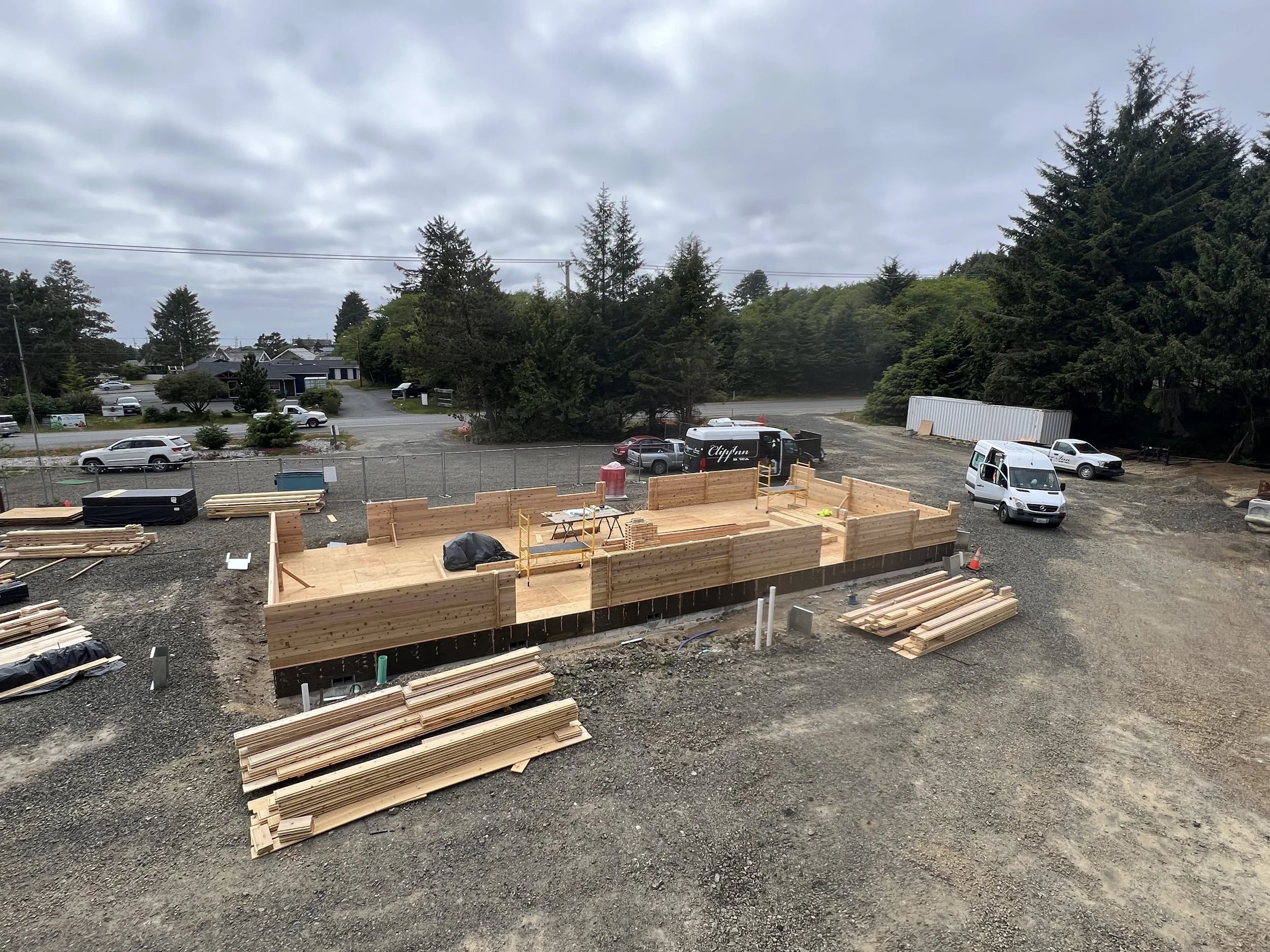 Construction site with a wooden foundation, several stacks of lumber, vehicles parked on the gravel, and a cloudy sky overhead.