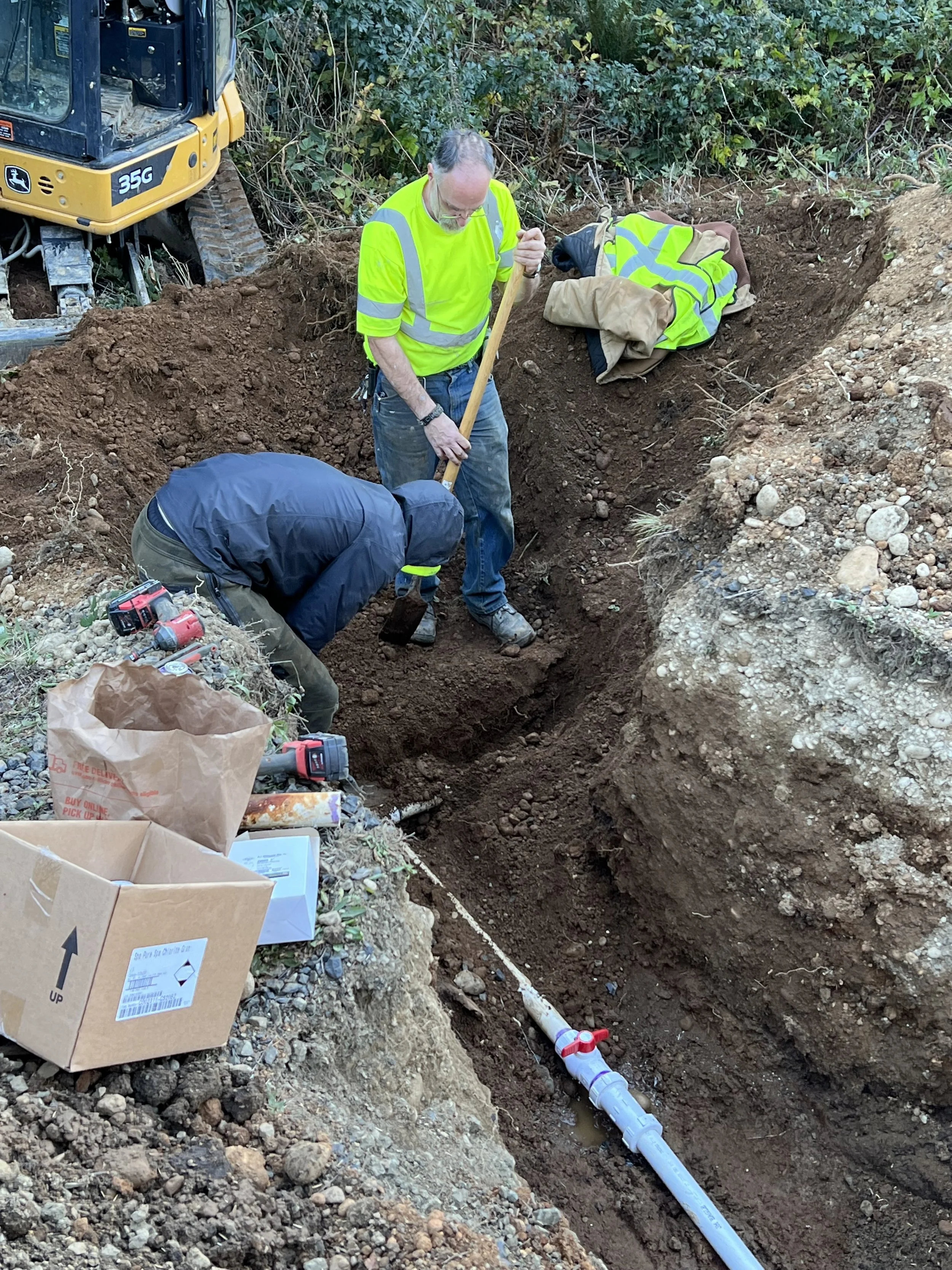 Two workers are installing underground pipes in a trench. One worker is standing and holding a shovel, while the other is crouching, working on the pipe. There is construction equipment, tools, and a cardboard box nearby, with dense greenery in the b