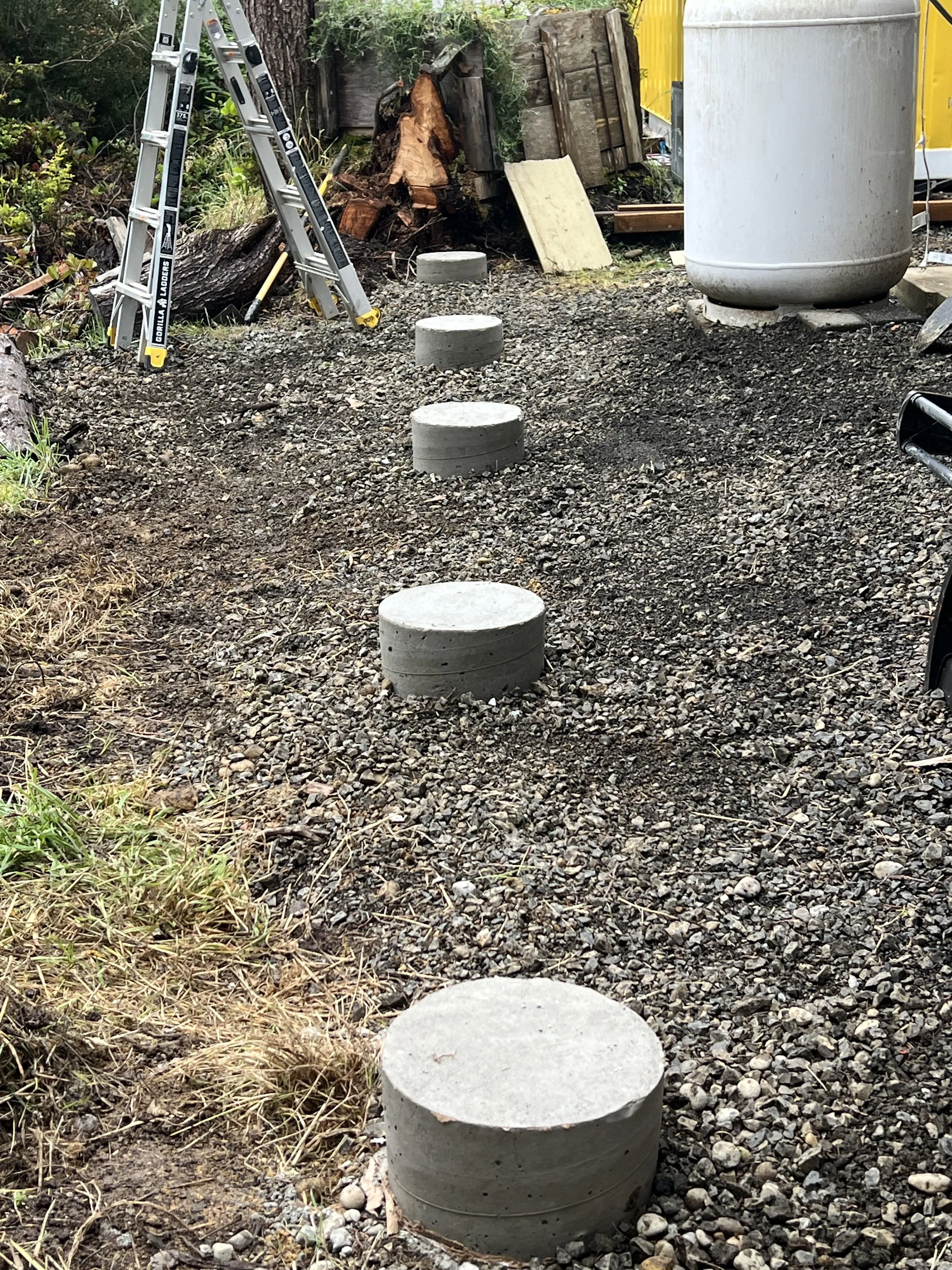 A row of four concrete steps emerging from dirt on a construction site, with a ladder, wooden planks, and trees in the background.