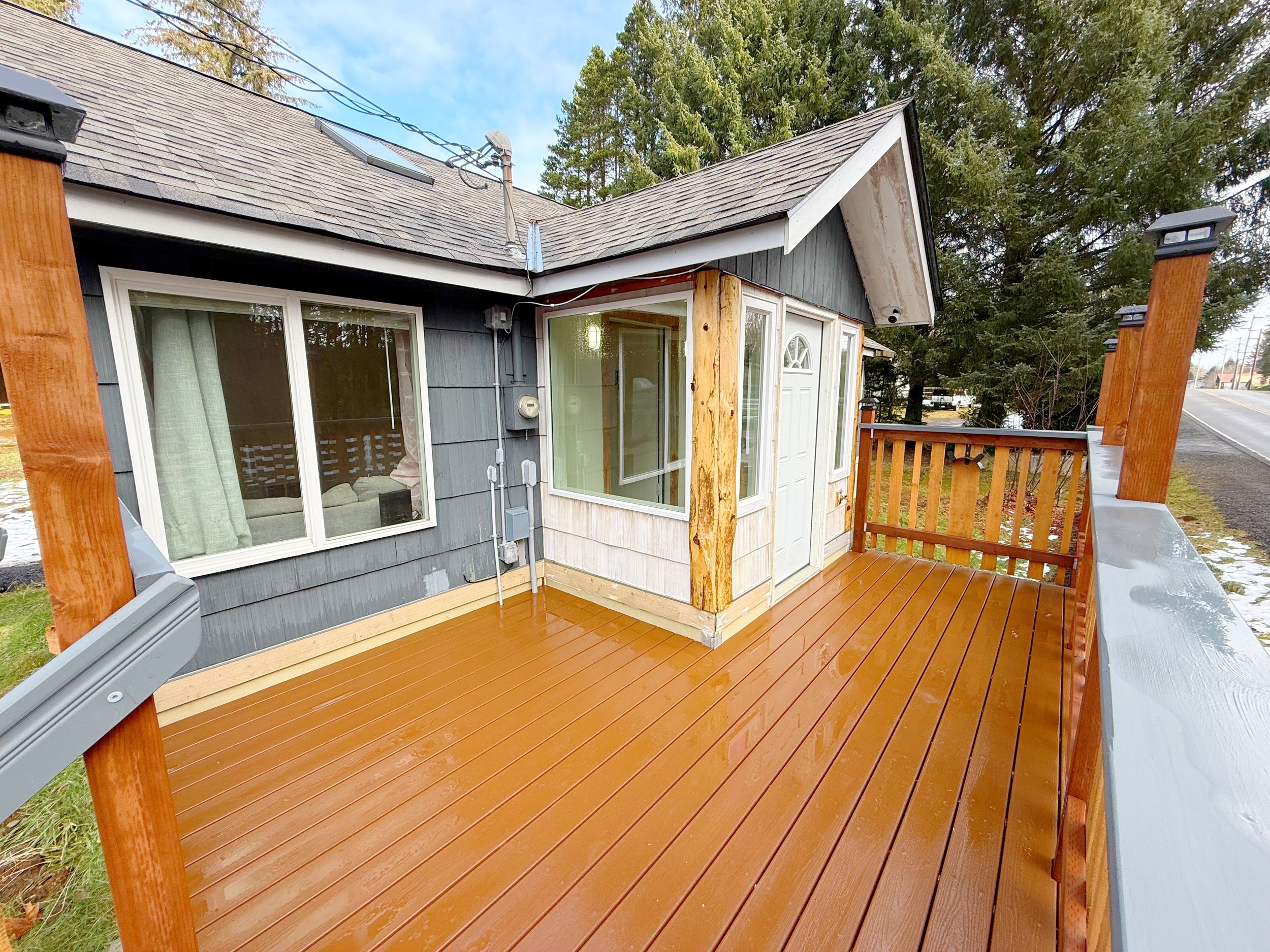 A wooden deck attached to a house, with wooden posts and a railing. The house has dark gray siding, a window with light-colored curtains, and a door with a glass panel. The roof has gray shingles, and there are electrical wires on the peak. Tall tree
