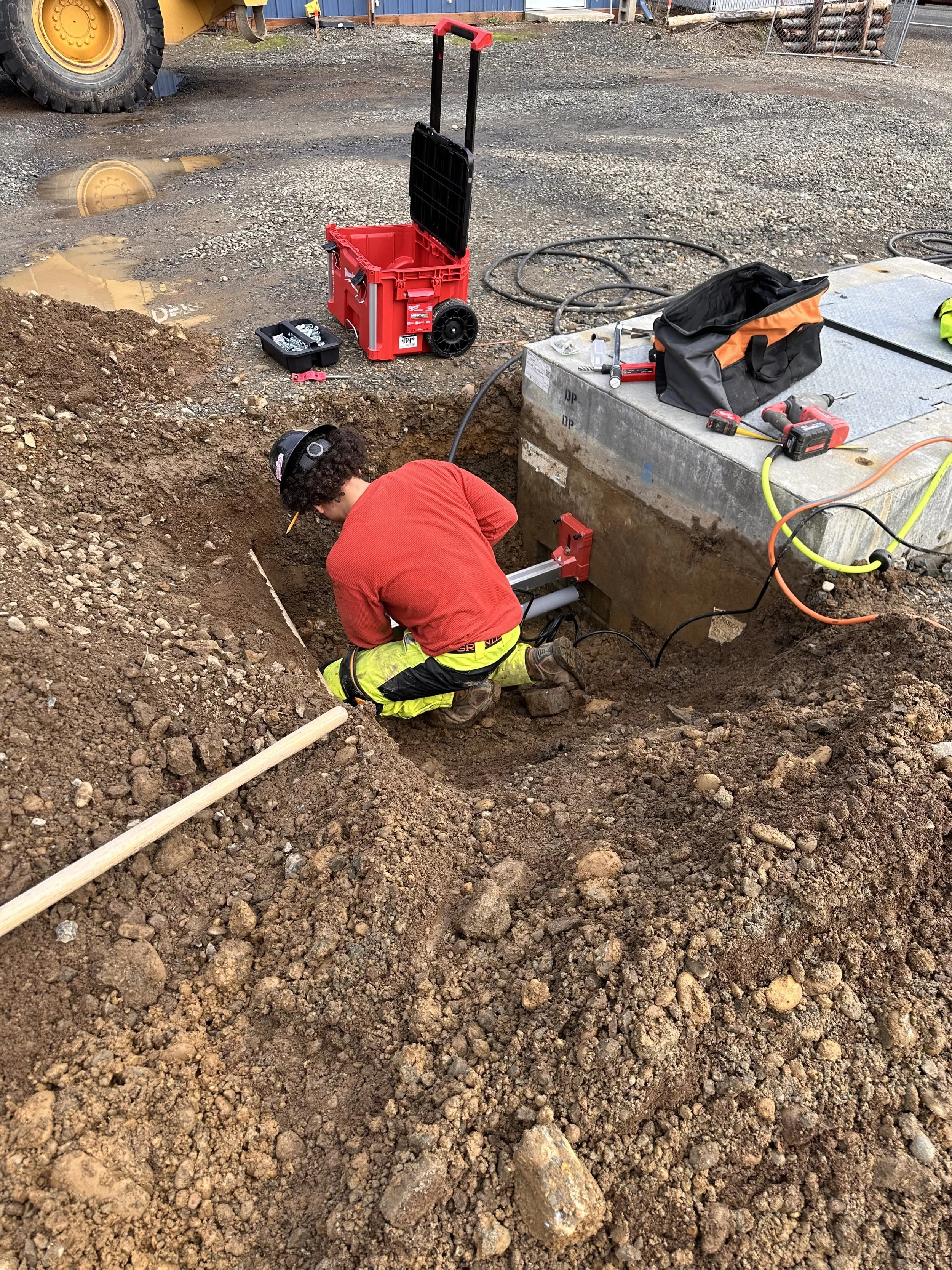 A construction worker in yellow safety pants and a red shirt working in a hole in the ground, surrounded by dirt and construction tools, with an orange and black bag and a metal box nearby, at a construction site with machinery and materials in the b