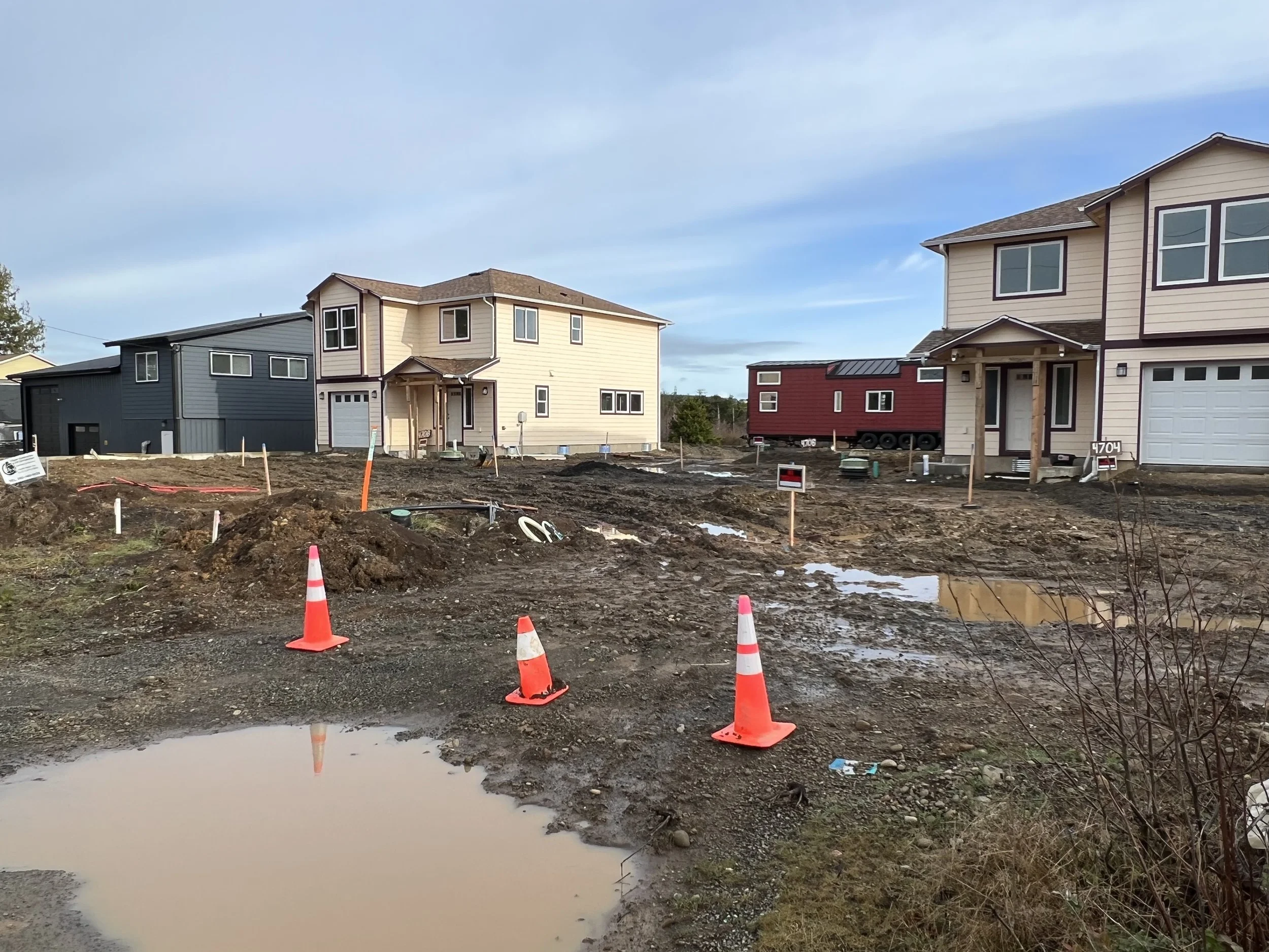 A construction site with new houses and orange safety cones, muddy ground with puddles, and a partly cloudy sky.