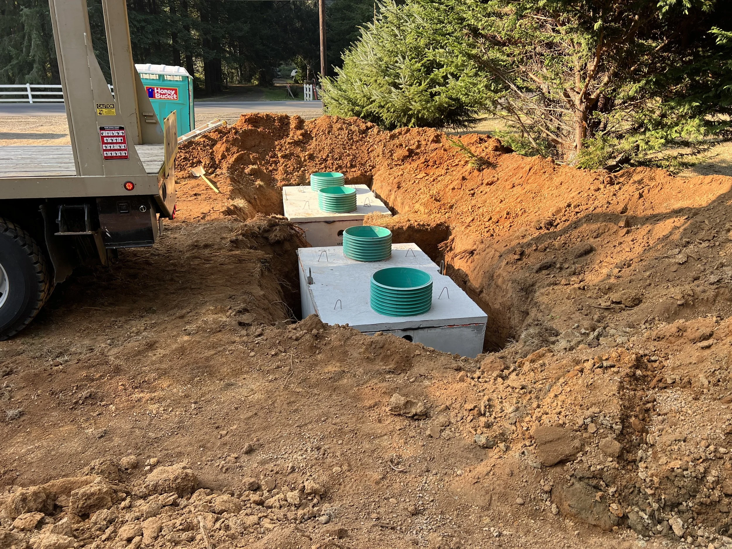 Construction site with dug trench, concrete bases with green pipes, dirt mounds, trees, and a portable toilet in the background.