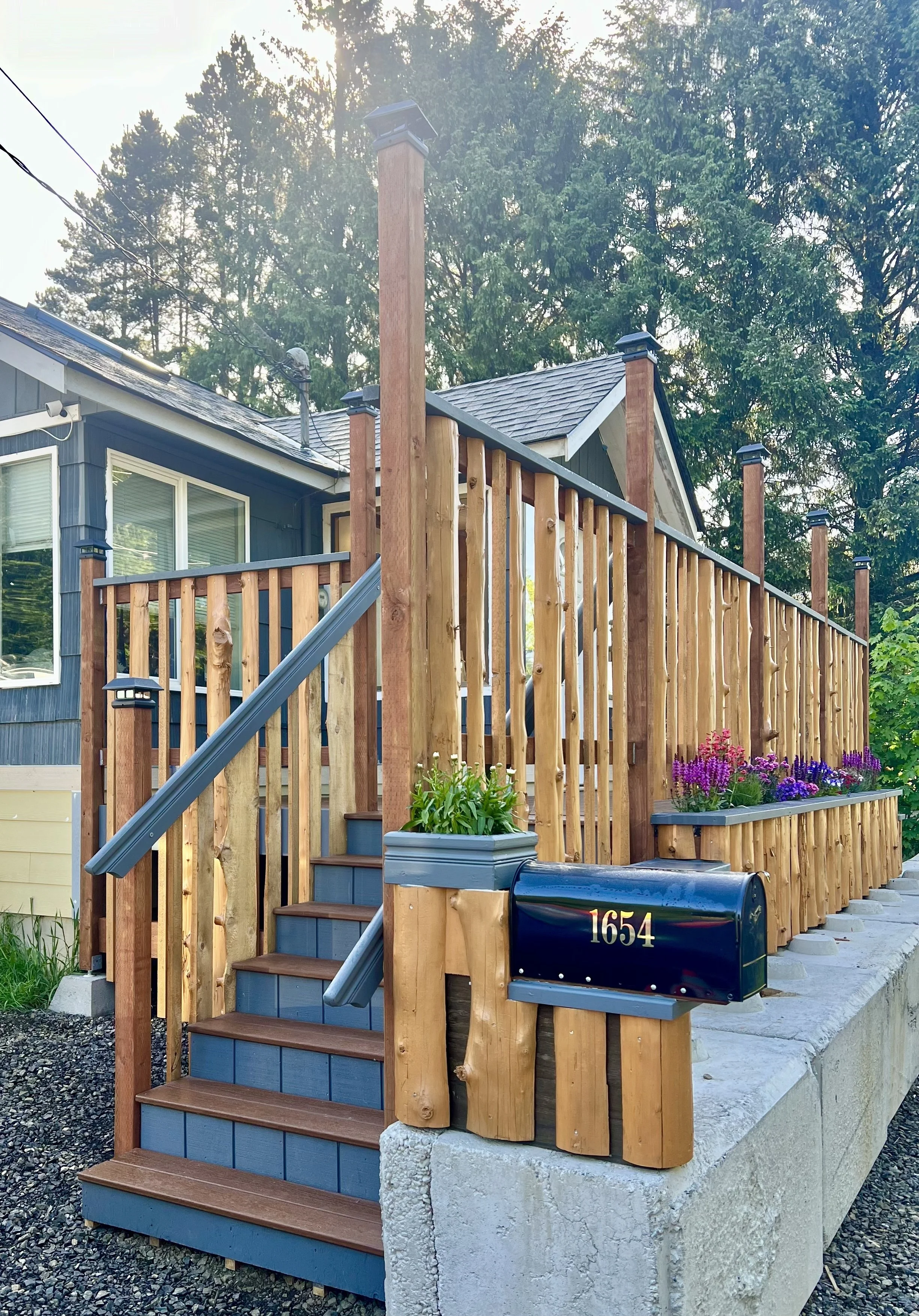 A house with a wooden deck, stairs, and railing in front and a dark blue exterior, with trees in the background. There are flowers and a mailbox with the number 1654.