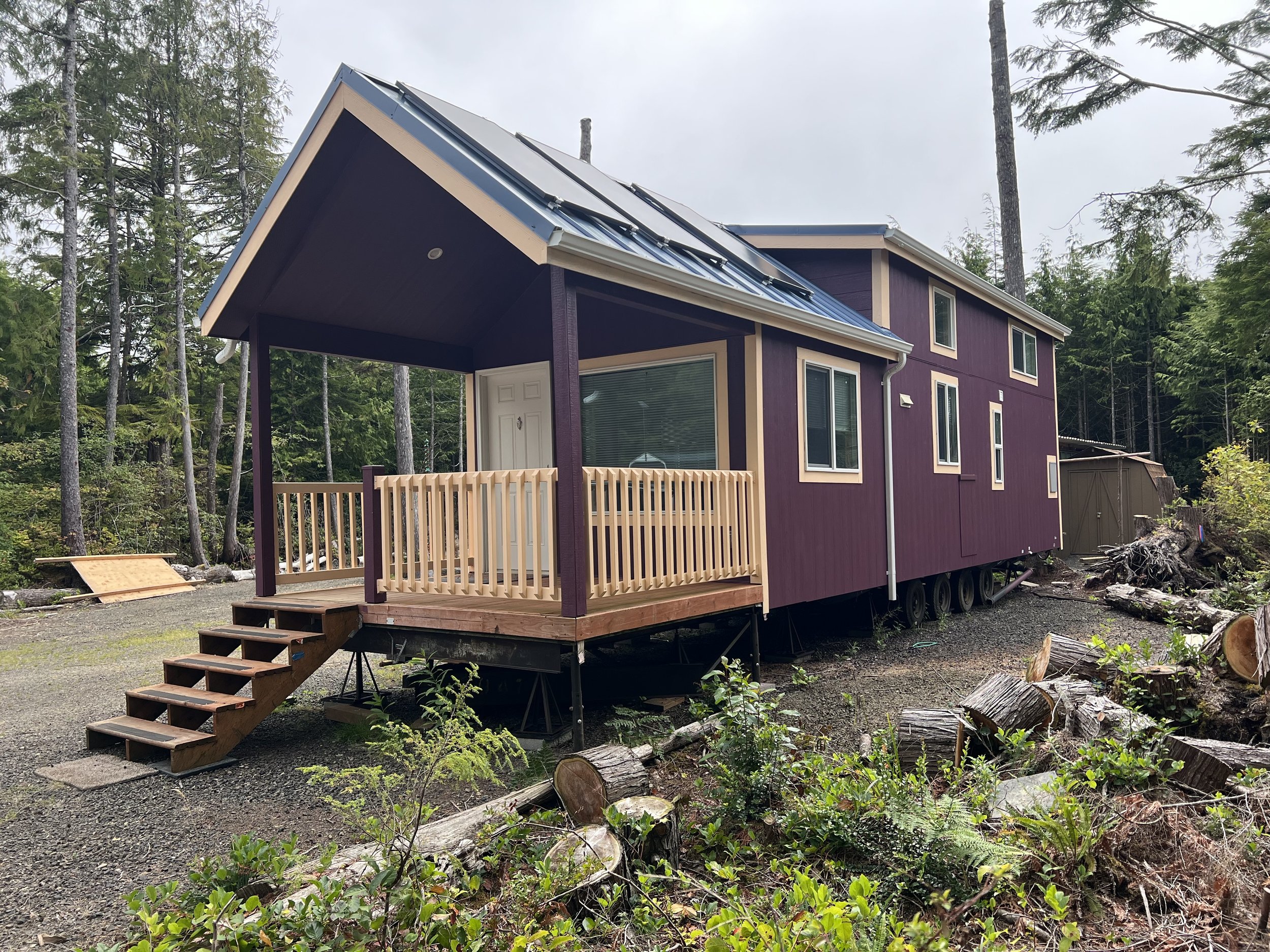 A purple house with beige trim is under construction in a forested area. The house is elevated on a trailer with stairs leading up to the front door. The background includes tall evergreen trees and a cloudy sky.