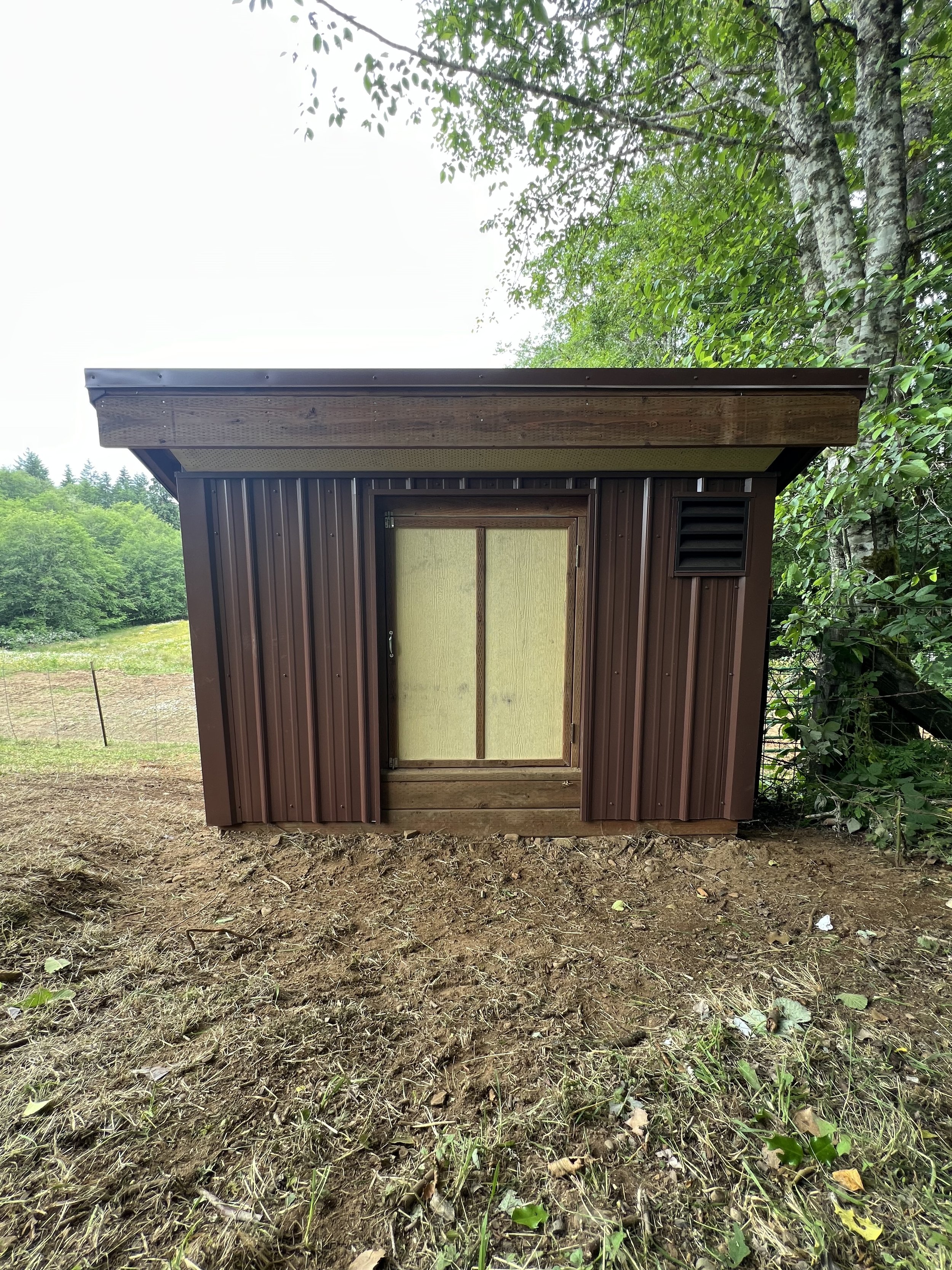 Small brown shed with a metal roof and a sliding door, surrounded by trees and grass, in a rural outdoor setting.
