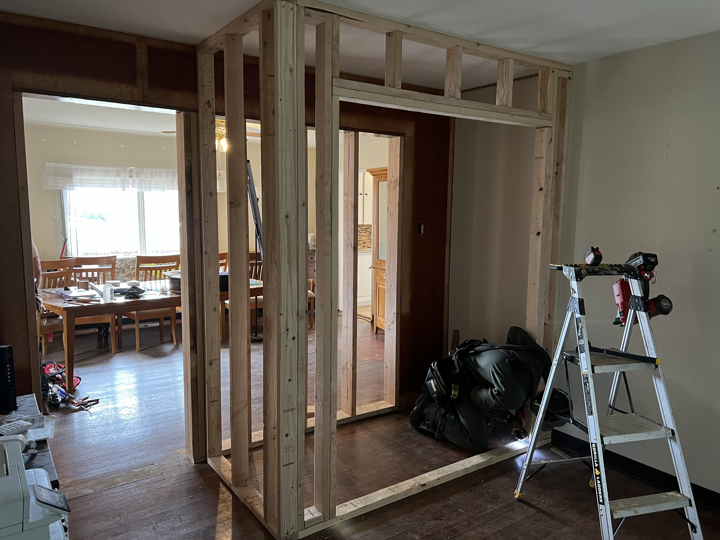 Interior view of a house under renovation, showing a wooden framing structure and construction tools, including a step ladder and tool bags.