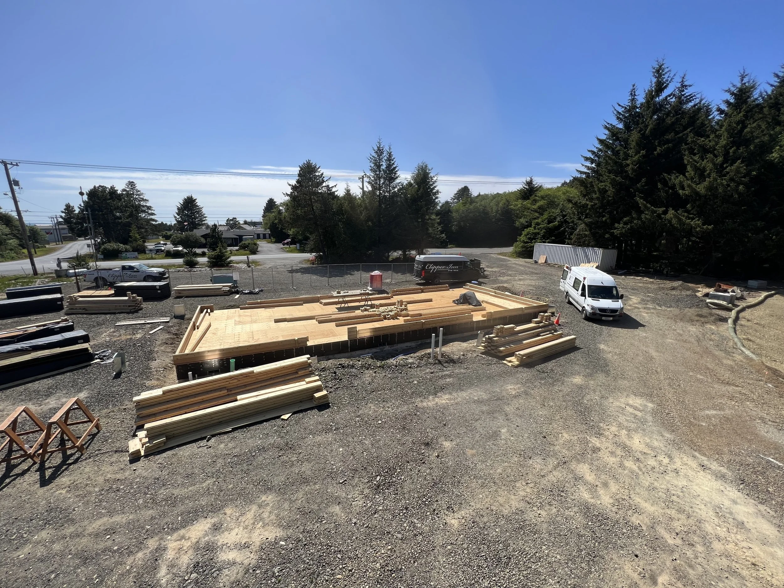 Construction site with wooden framing, construction materials, and vehicles, with trees and a clear blue sky in the background.