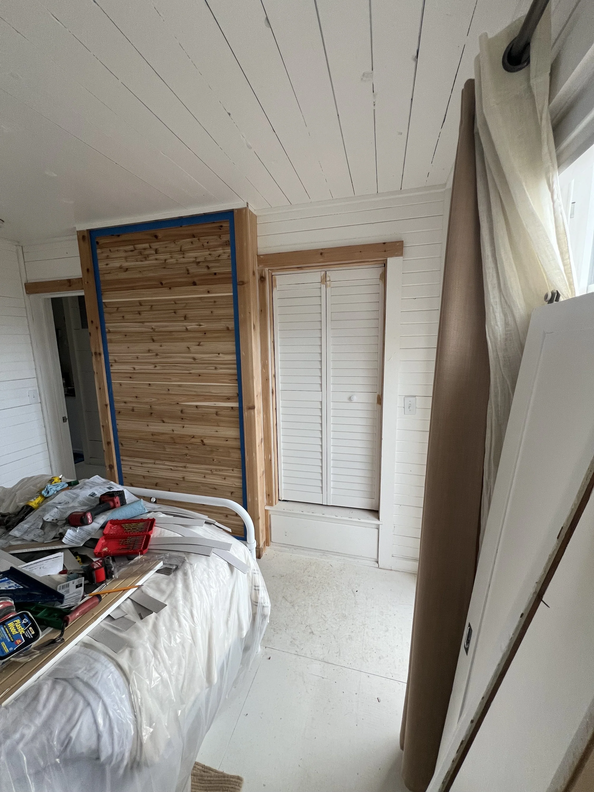 Part of a bedroom under renovation with a bed covered in tools, wood paneling on wall, closets, curtains, and unfinished trim.