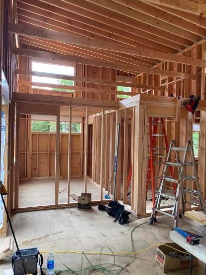 Interior view of a residential construction site with wooden framing, partially built walls, and construction tools and ladders.
