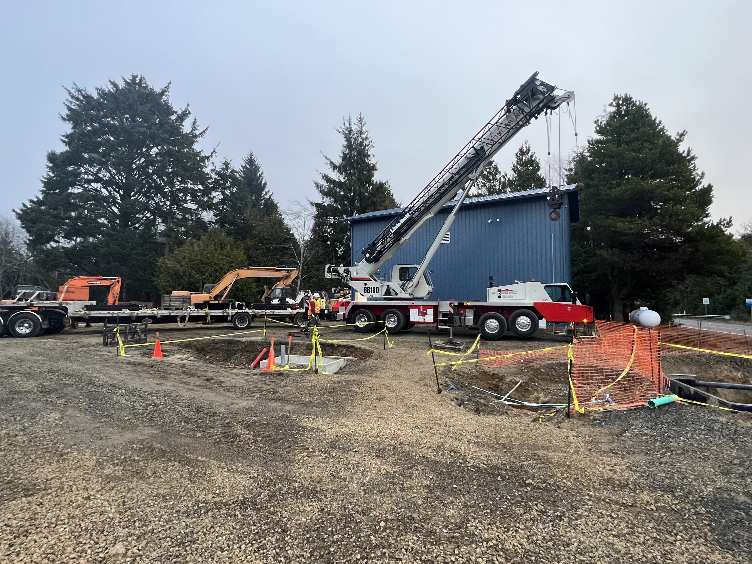Construction site with excavation area, yellow caution tape, orange traffic cone, a crane truck, and workers in safety gear, alongside equipment and vehicles, with trees in the background.