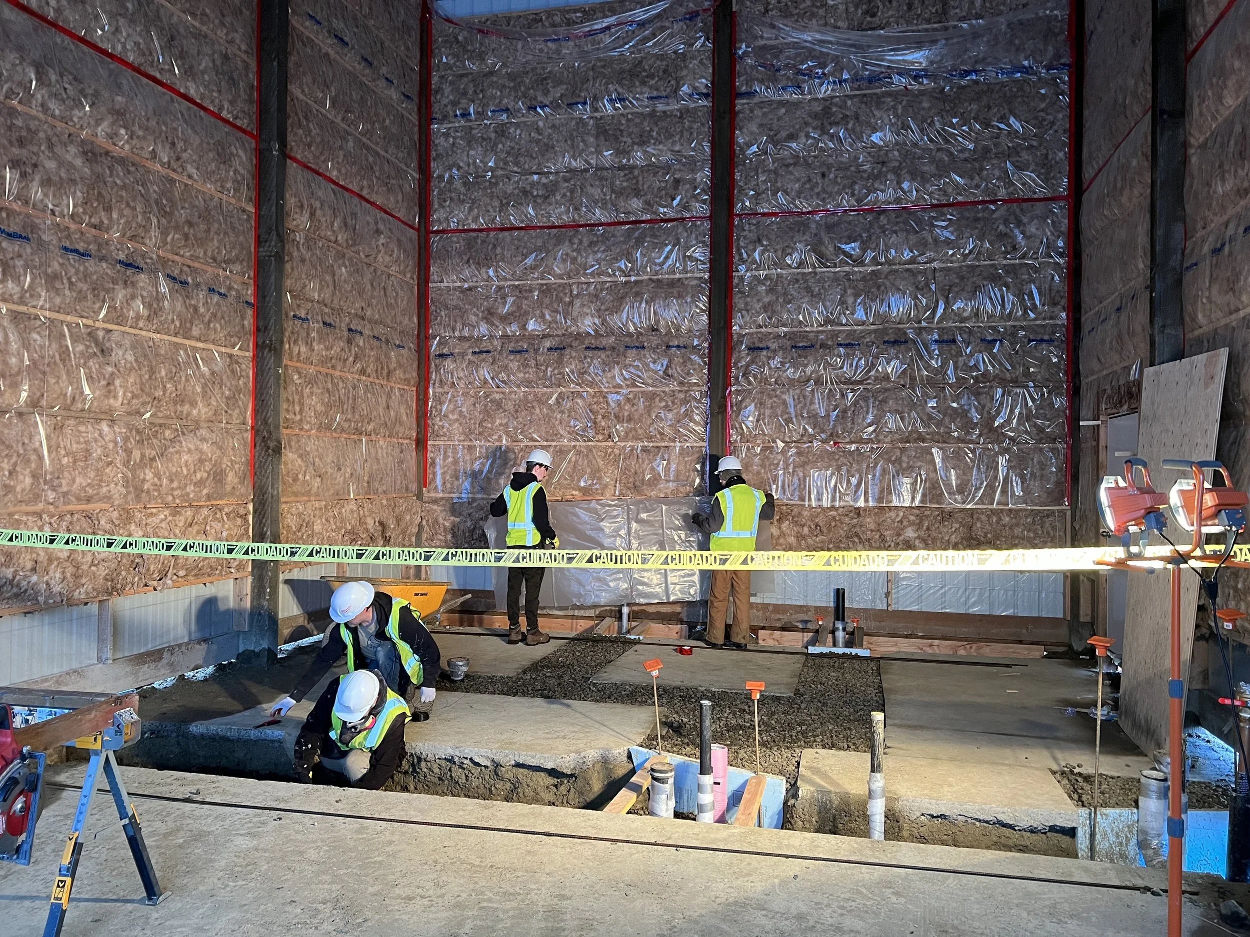 Construction workers wearing safety helmets and high-visibility vests working inside a building under construction, with wooden framing, insulation, and caution tape.