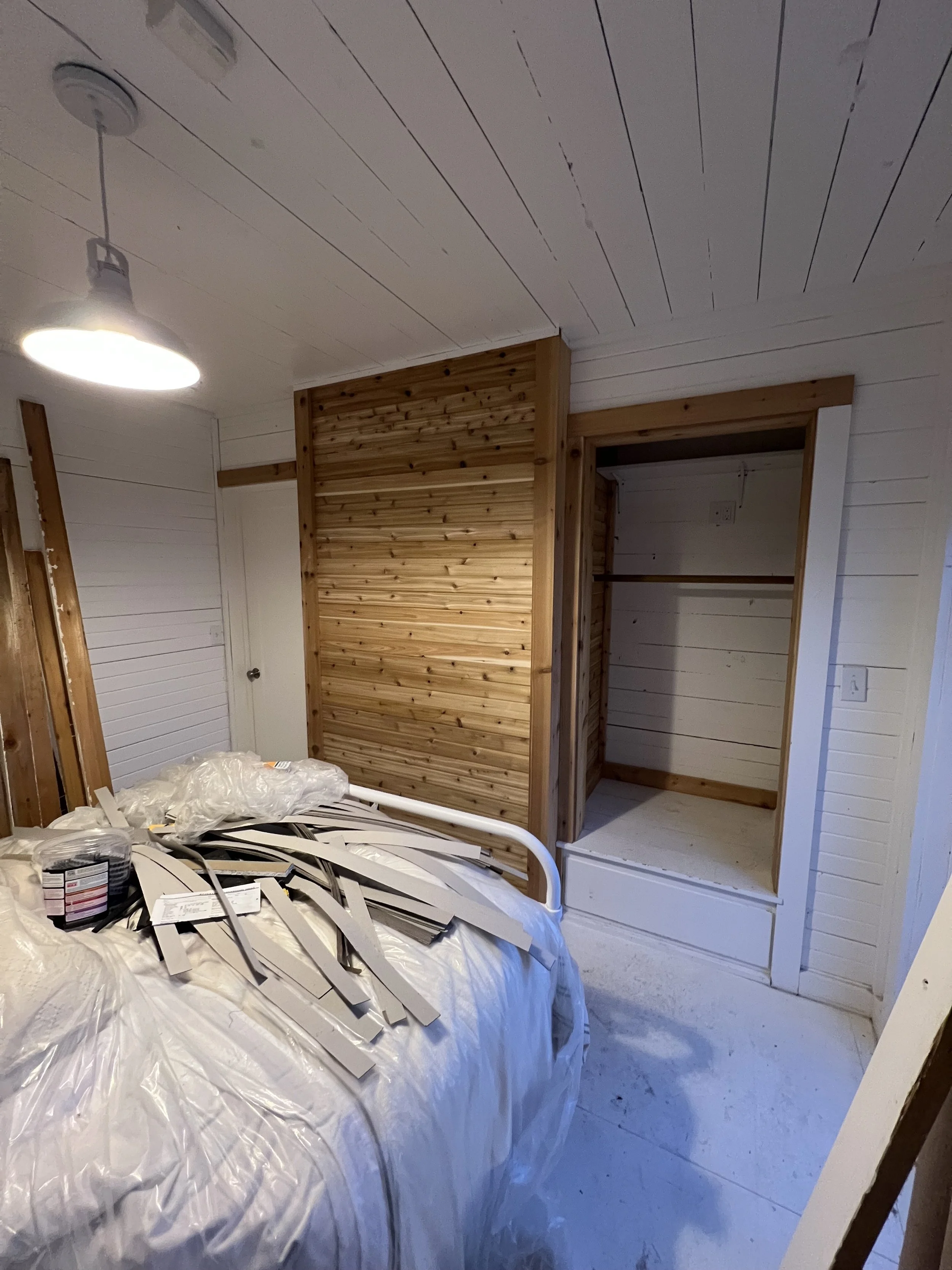 Interior of a room under renovation, with a partially assembled white wood closet frame and a pile of wiring and construction materials on a plastic-covered surface in the foreground. The ceiling and walls are painted white, with wood panel accents.