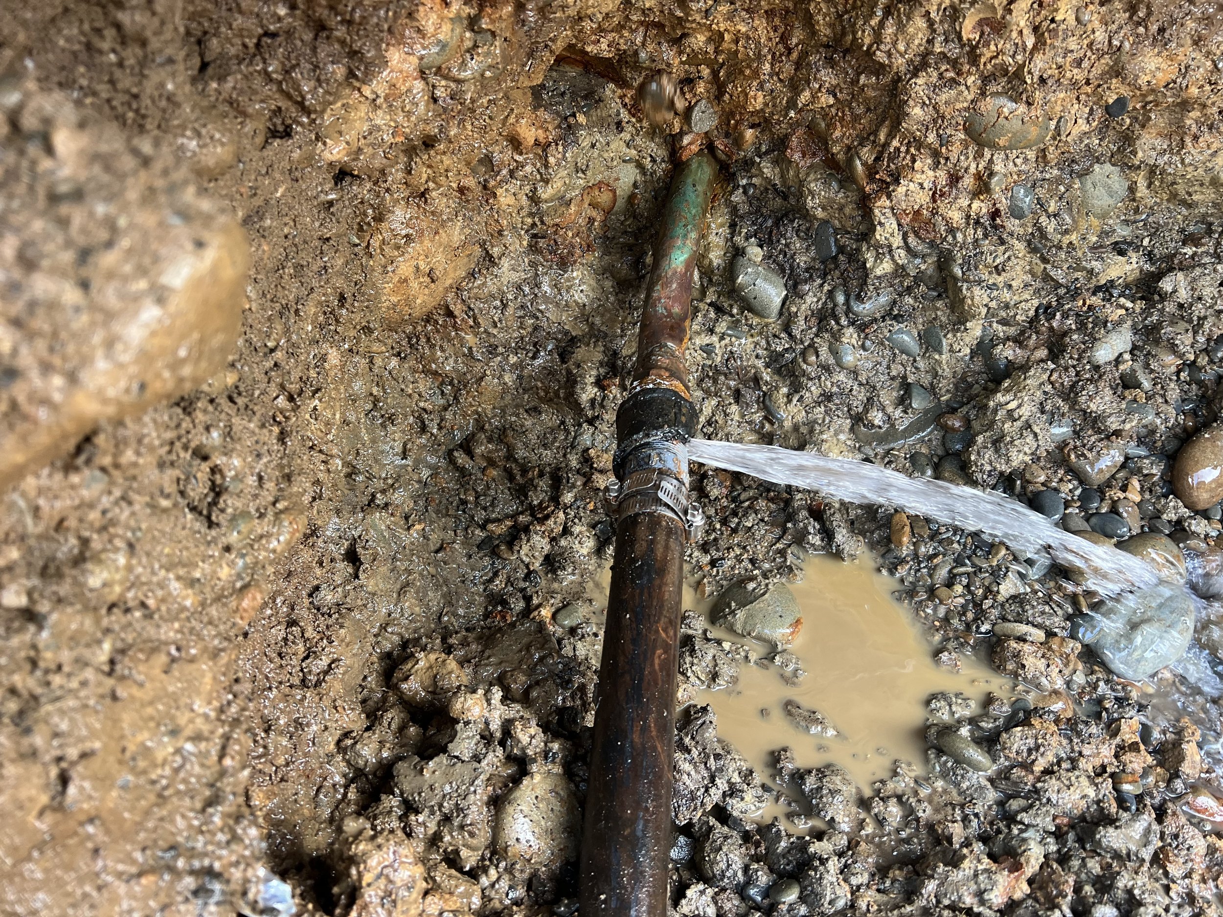 Close-up of a water pipe leaking water onto muddy ground surrounded by small rocks.